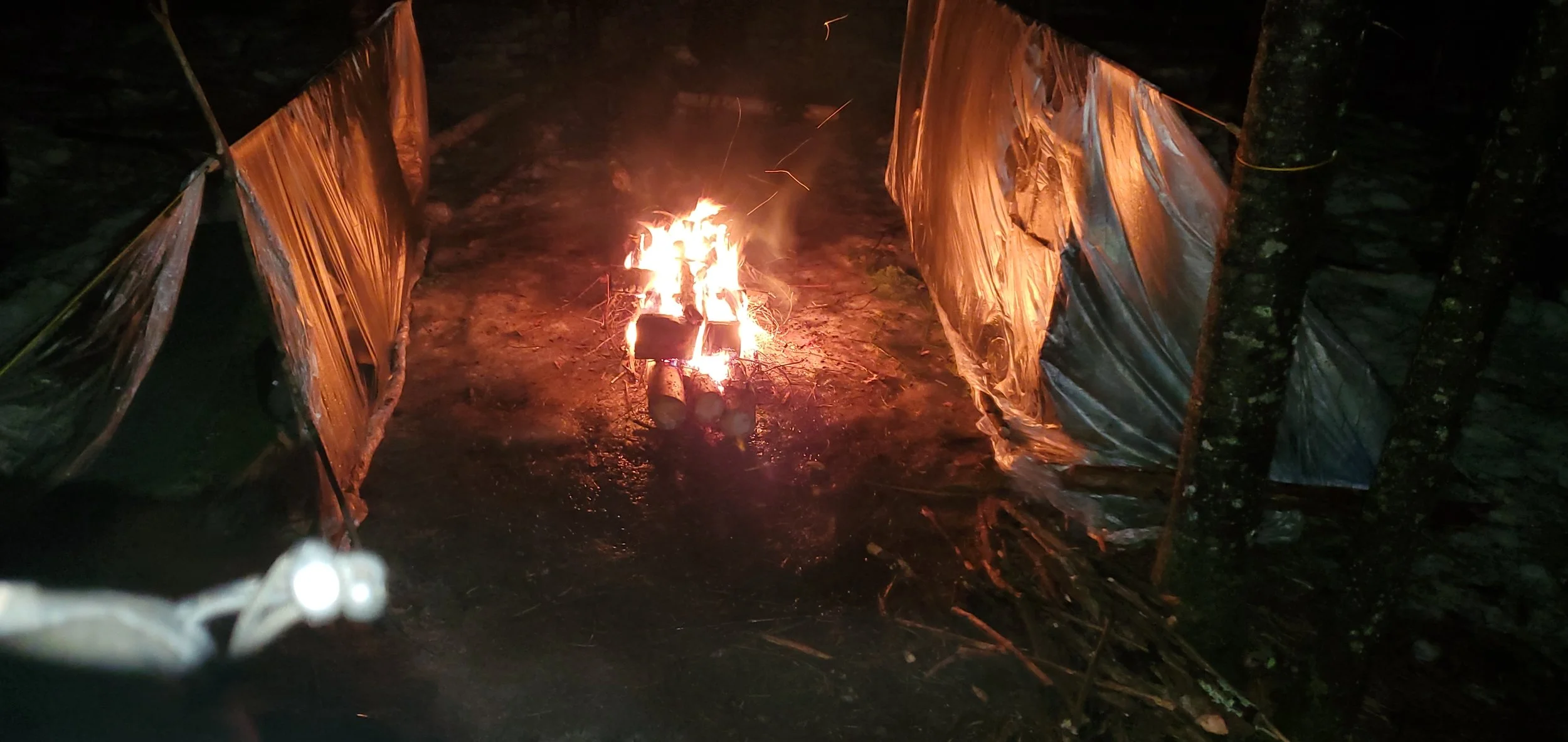 Campfire burning in a forest with a makeshift shelter made of reflective emergency blankets on either side.