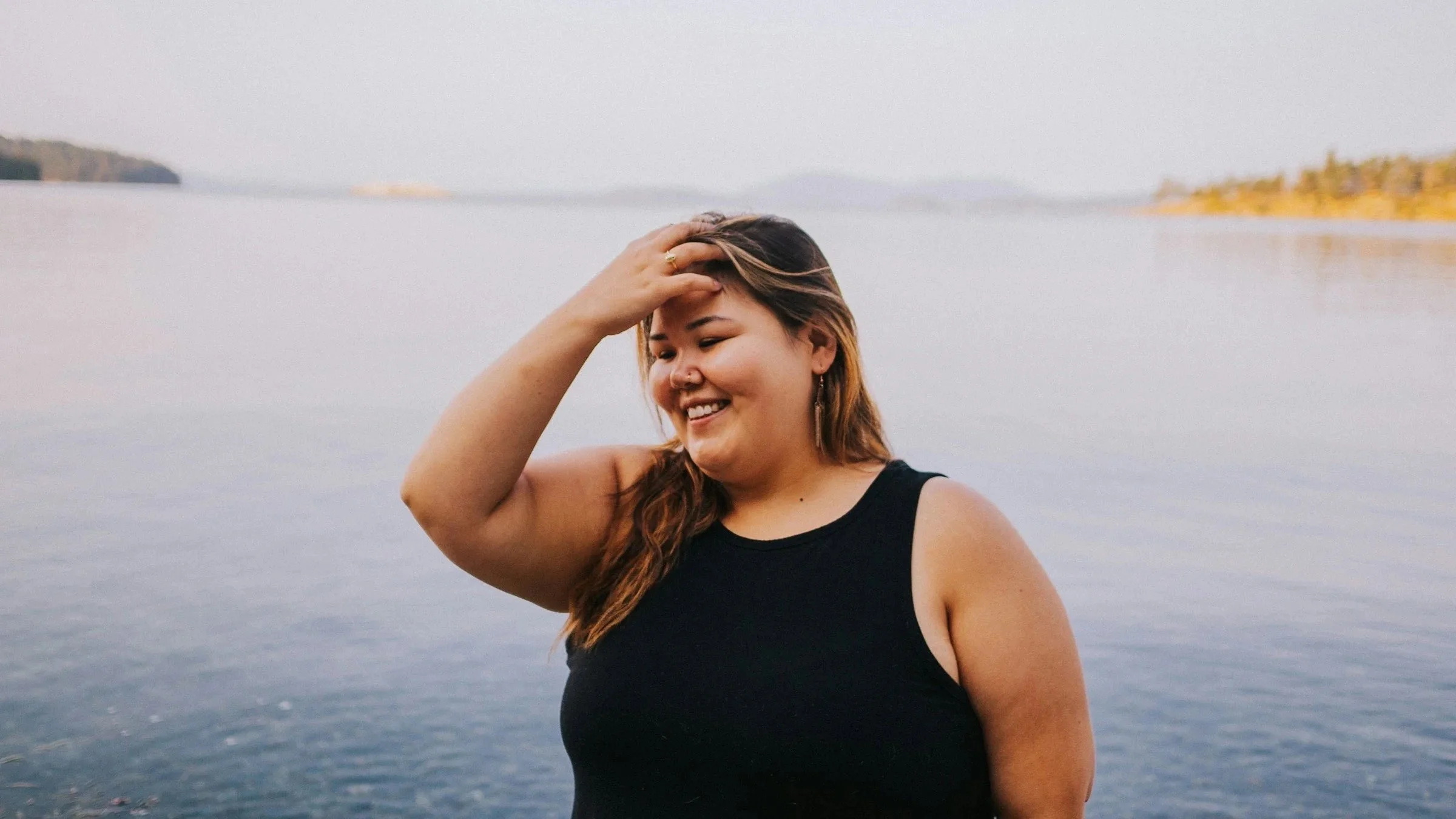 Woman with brown standing over a body of water smiling while toughing her hair