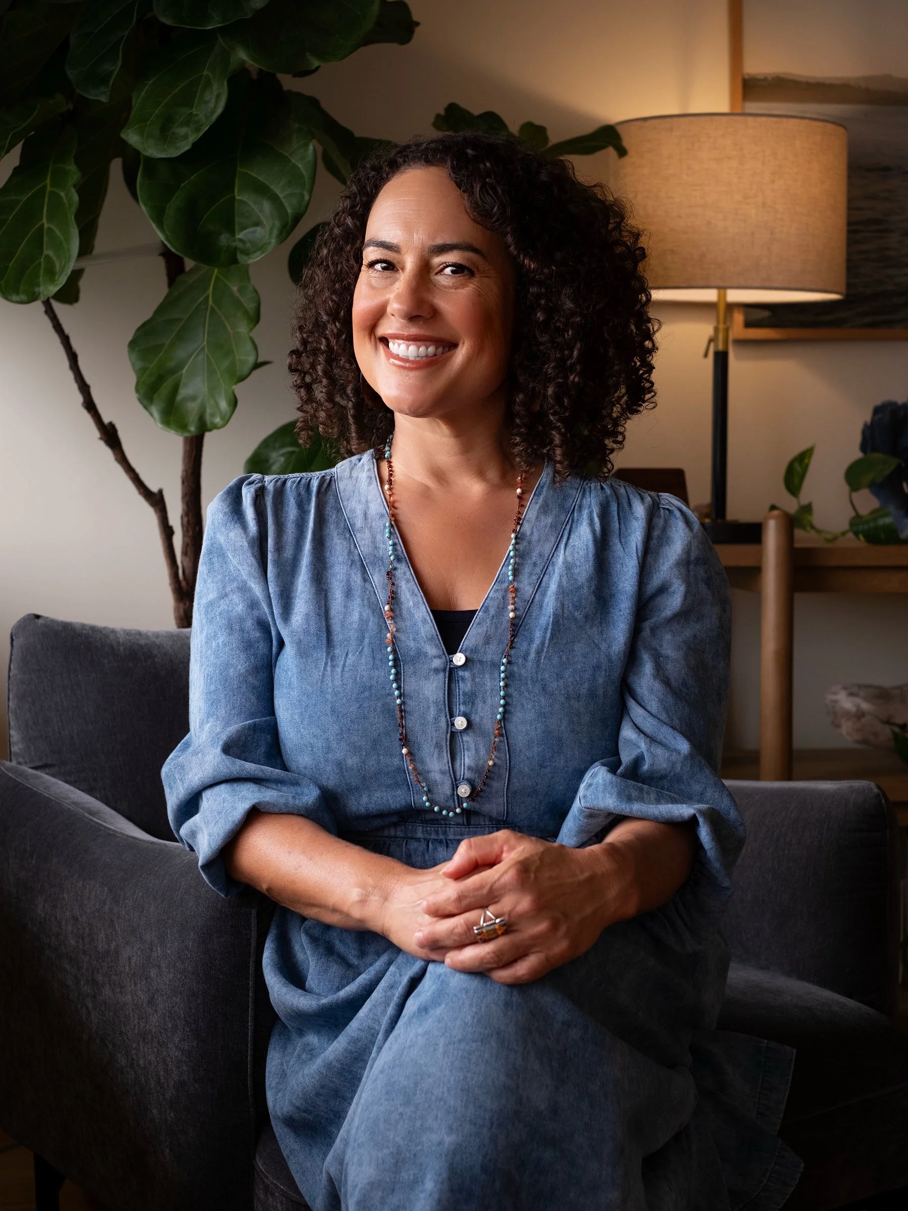 Woman with brown curly hair sitting in office with tree in background wearing denim blue dress