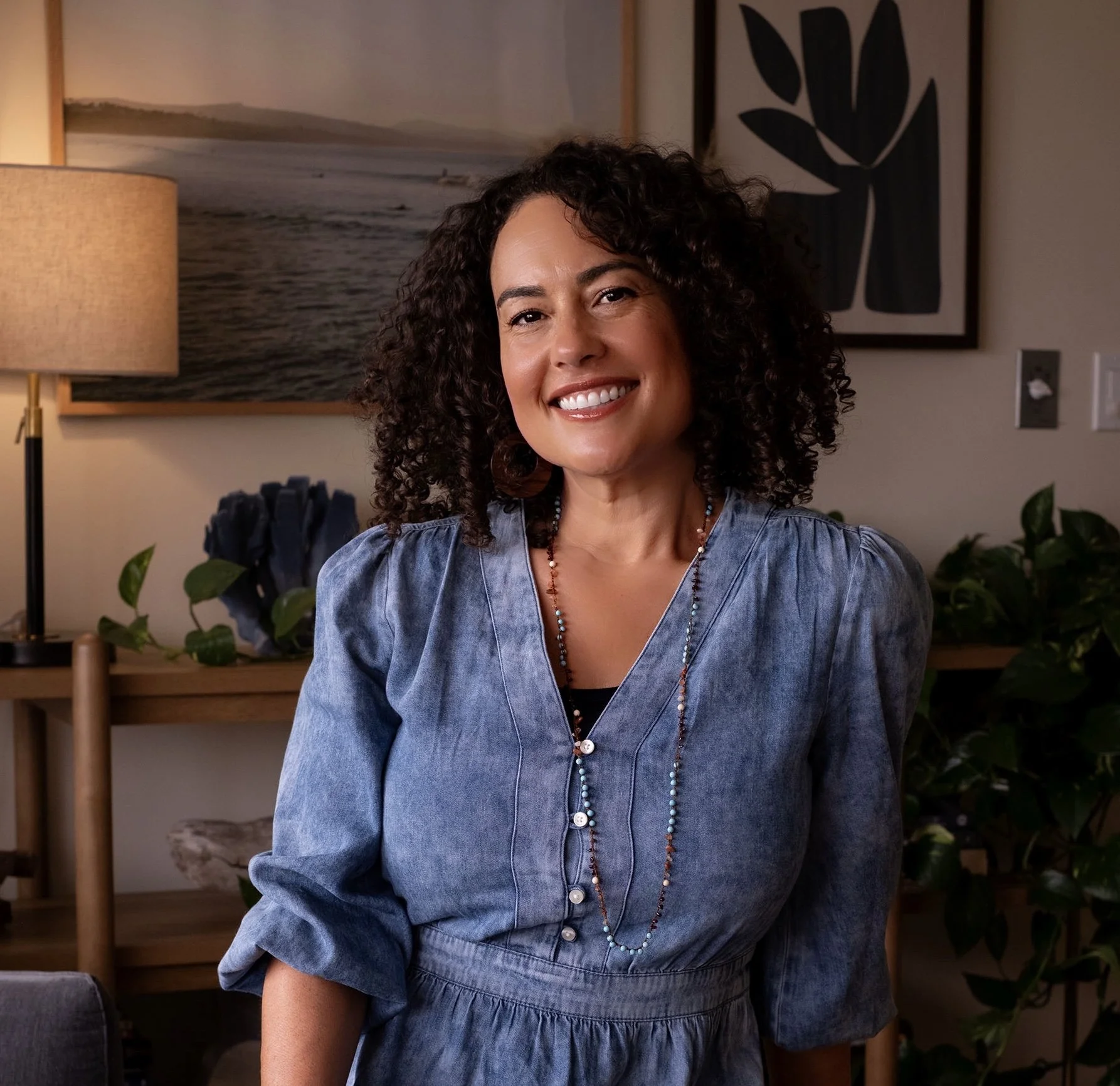 Smiling woman with brown curly hair wearing denim dress with turquoise beaded necklace sitting in office.