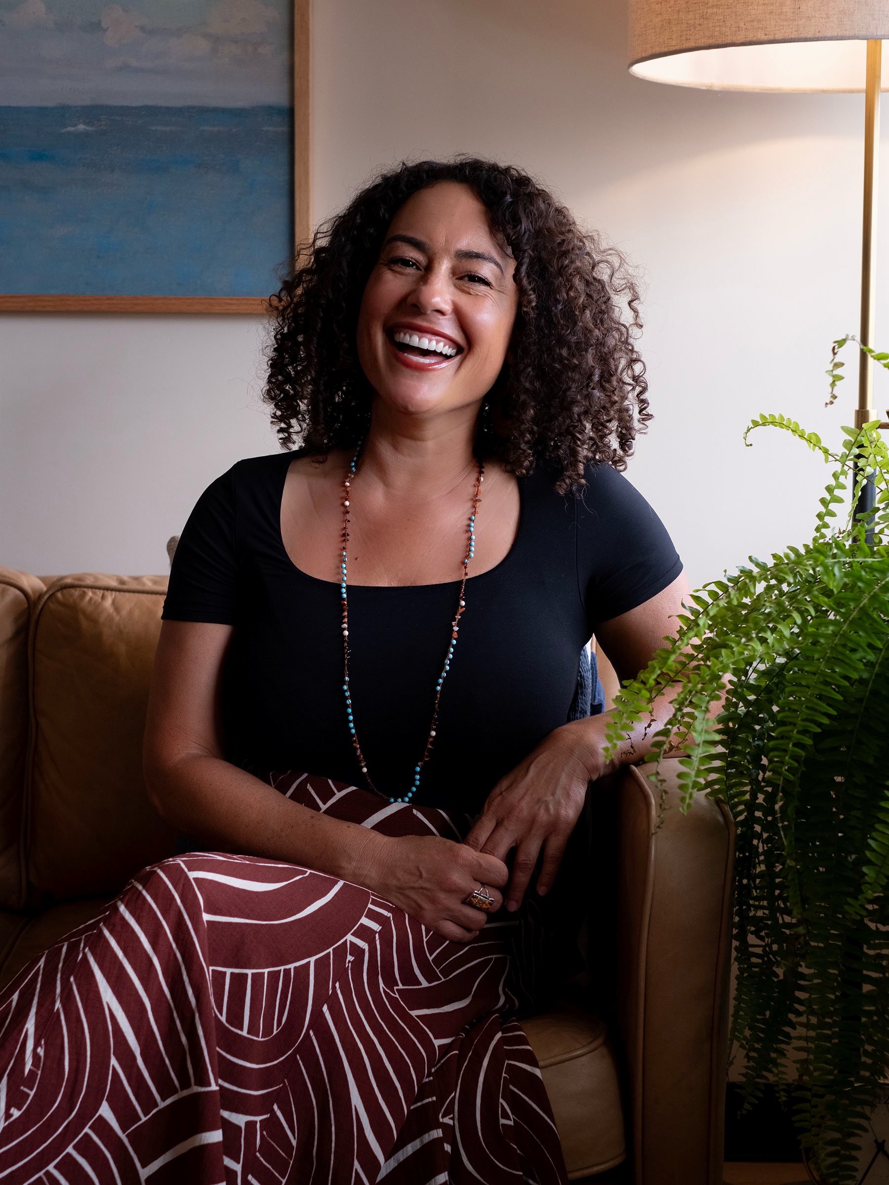 Female OCD Specialist with curly dark hair and a bright smile sitting in therapy office with black top and rust skirt.