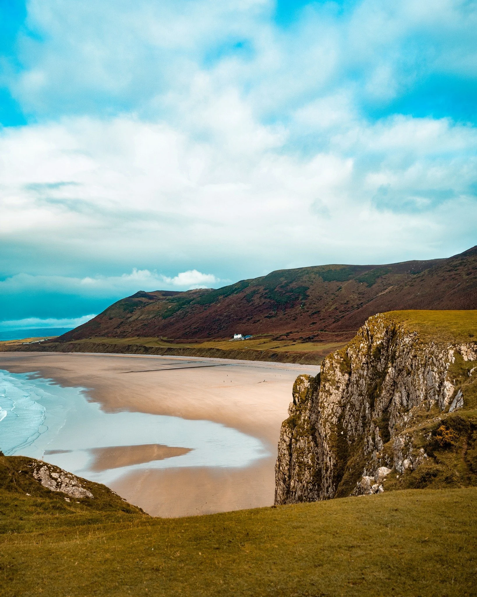 Coastal landscape with sandy beach, green hills, rocky cliffs, and blue sky.