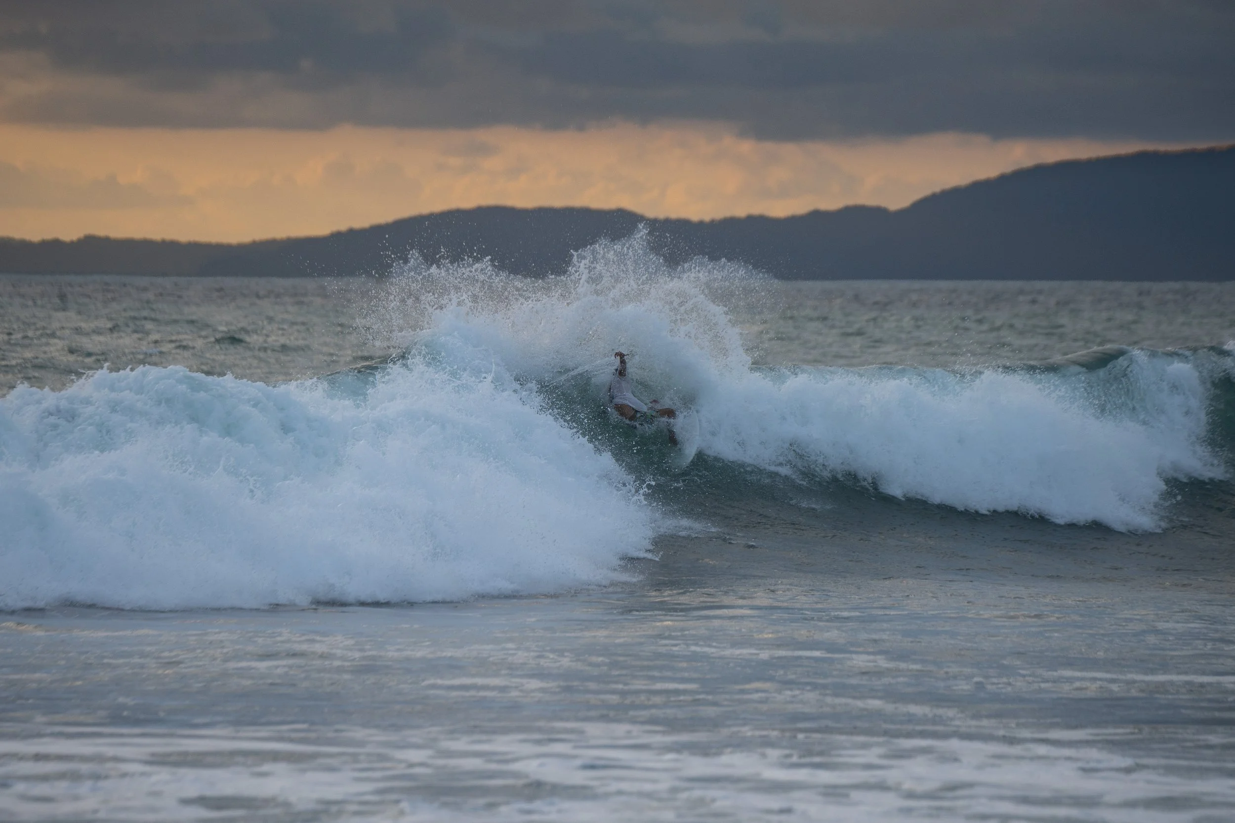 A surfer riding a wave in the ocean during sunset with mountains in the background.