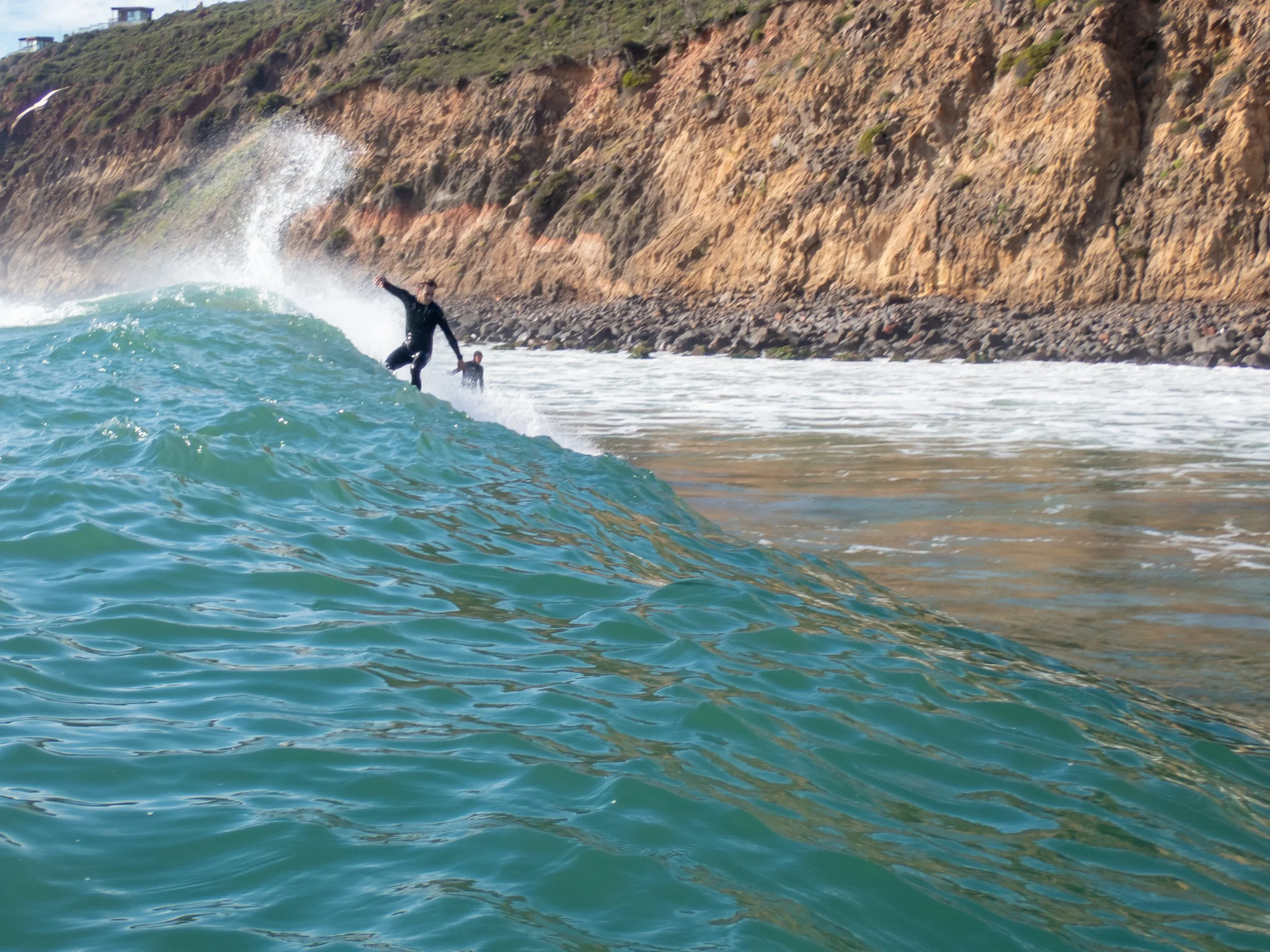 A person surfing on a wave near a rocky coastline with cliffs in the background.