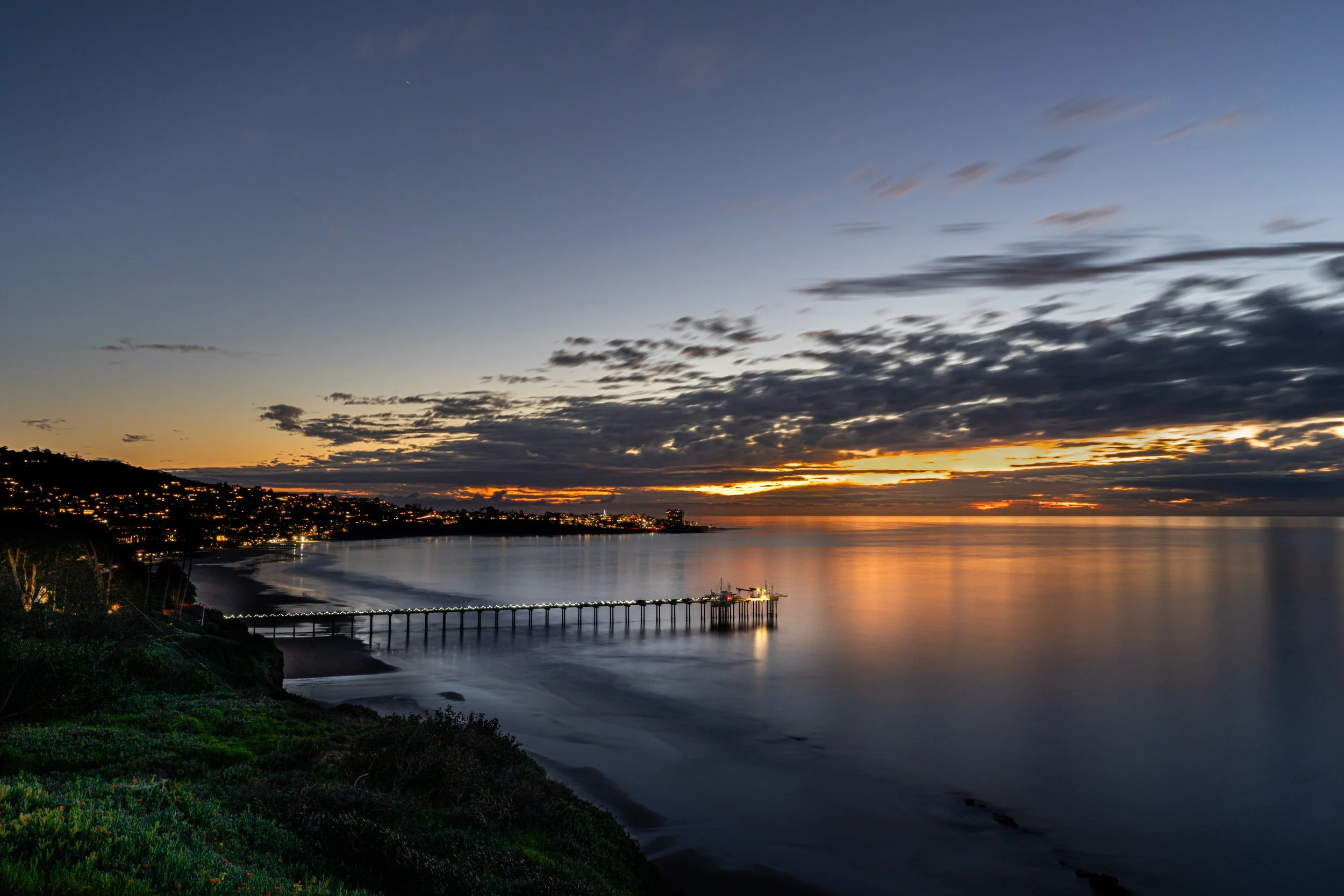 Sunset over a calm ocean with a pier extending into the water and a coastline with hills and houses with lights on.