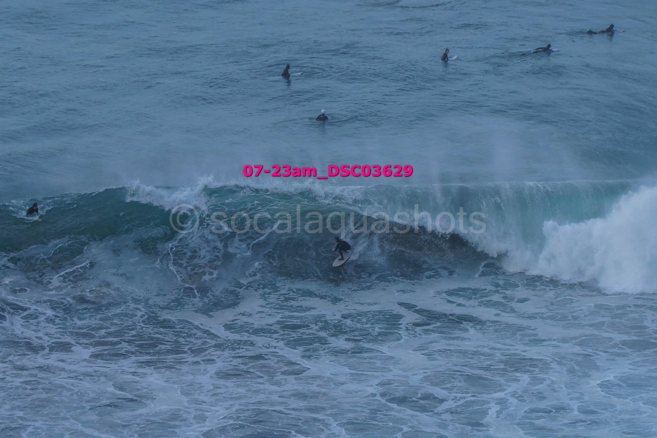 A surfer riding a wave in the ocean with multiple surfers in the background, some paddling and waiting for waves.