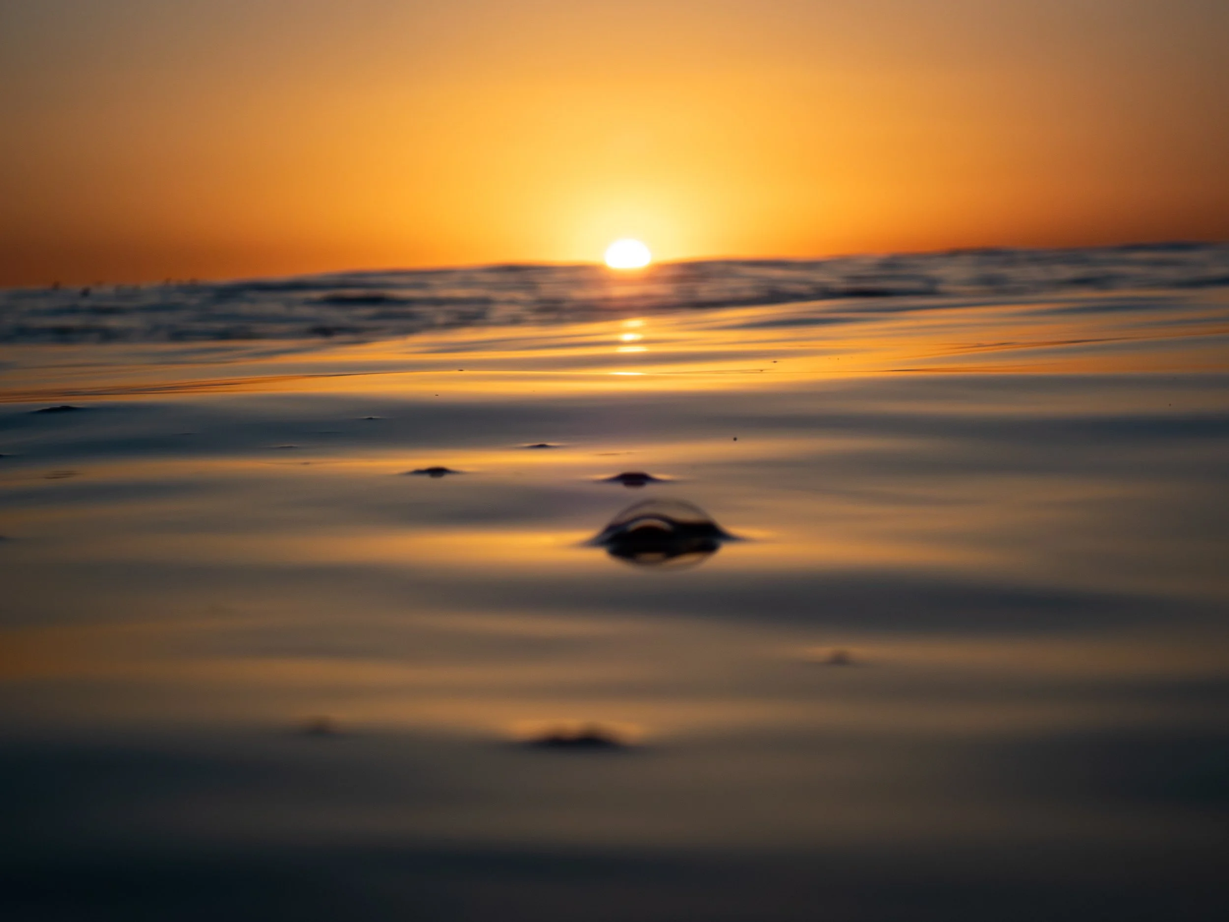 A low-angle view of a sunset over the ocean, with the sun near the horizon casting a golden glow, and gentle waves on the water surface.