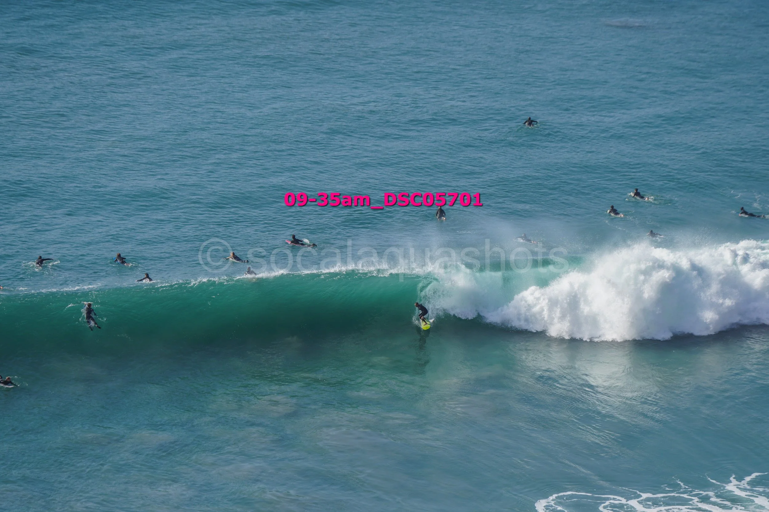 A surfer riding a large wave while multiple surfers wait in the water behind him in the ocean.