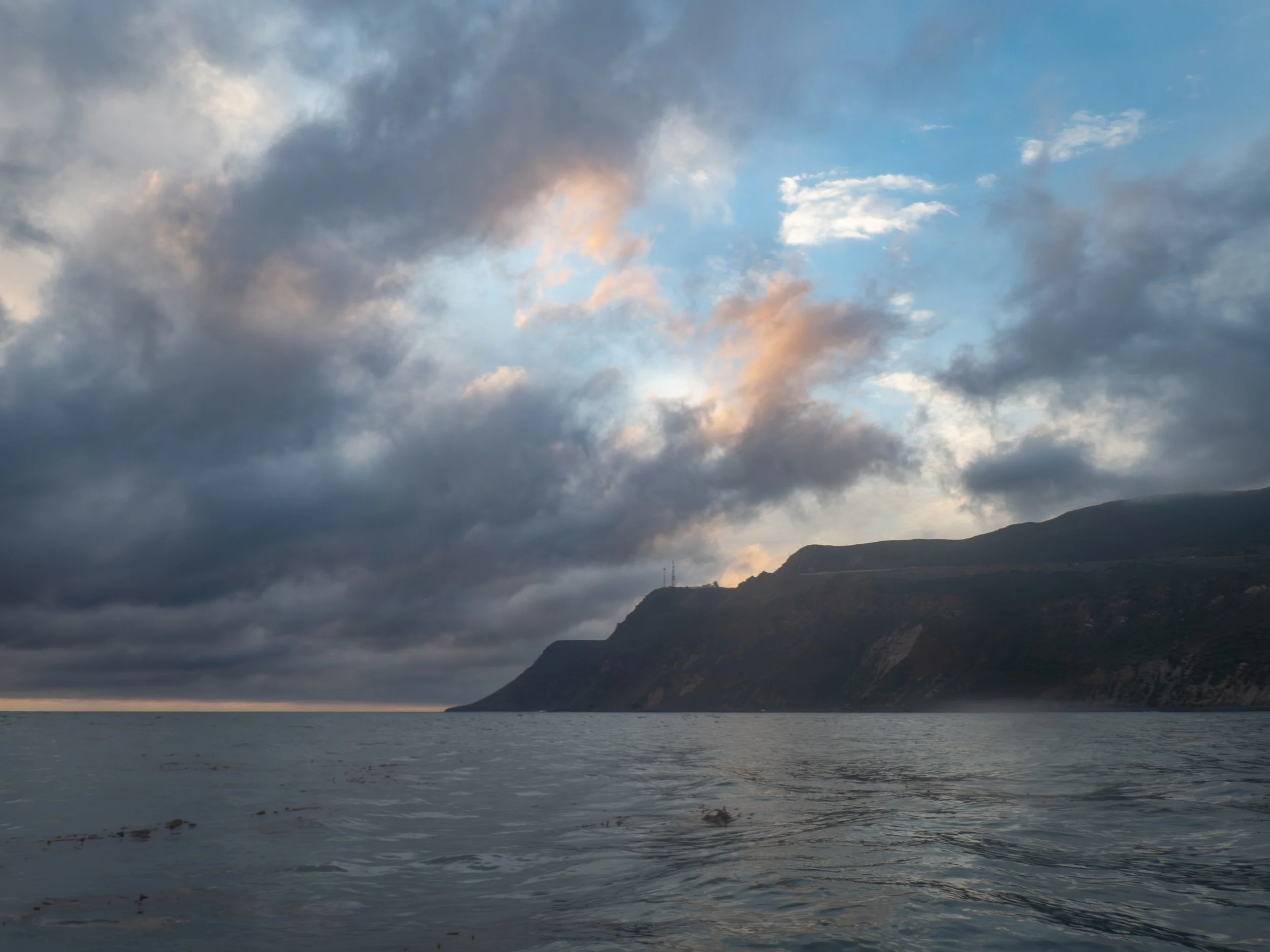 Overcast sky with clouds above a body of water and a hilly coastline in the distance.