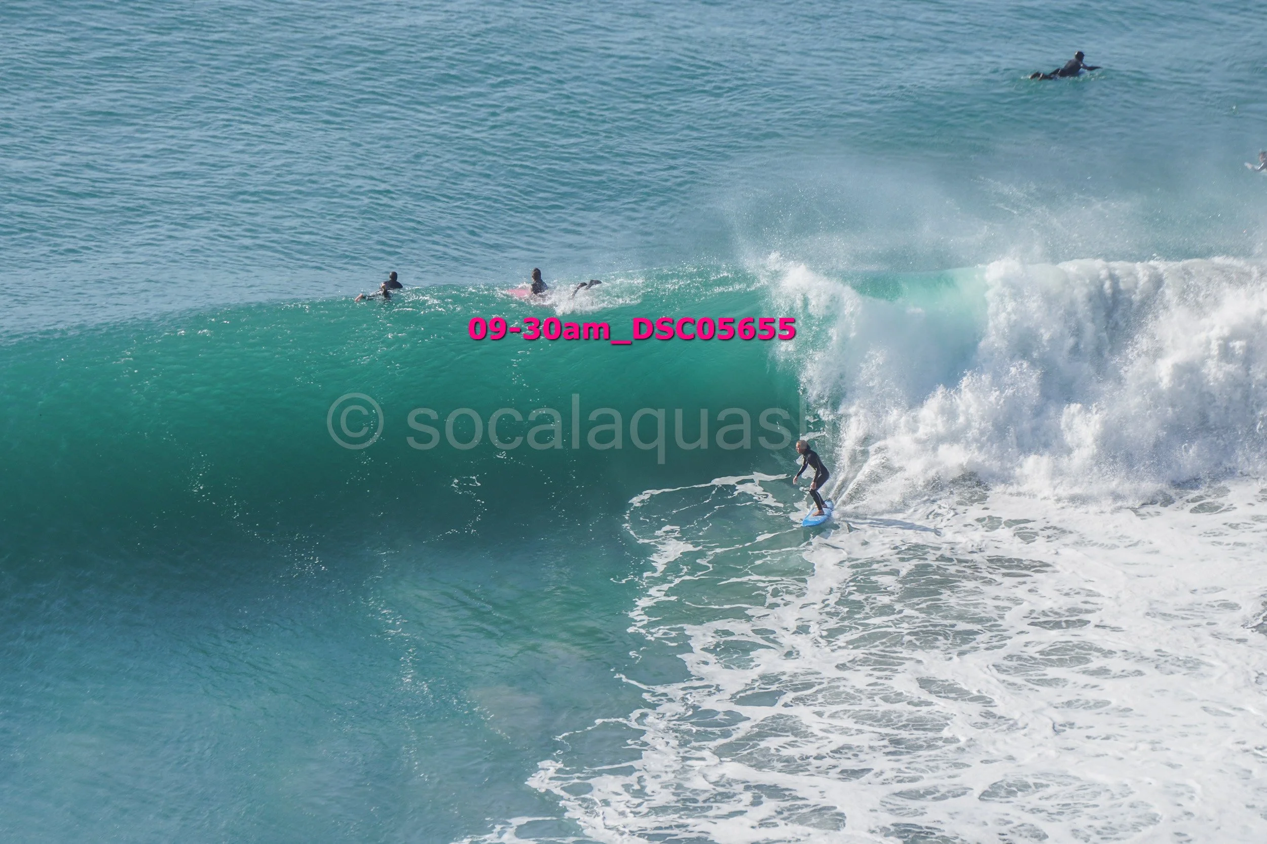 Surfer riding a large wave with several other surfers in the water nearby.