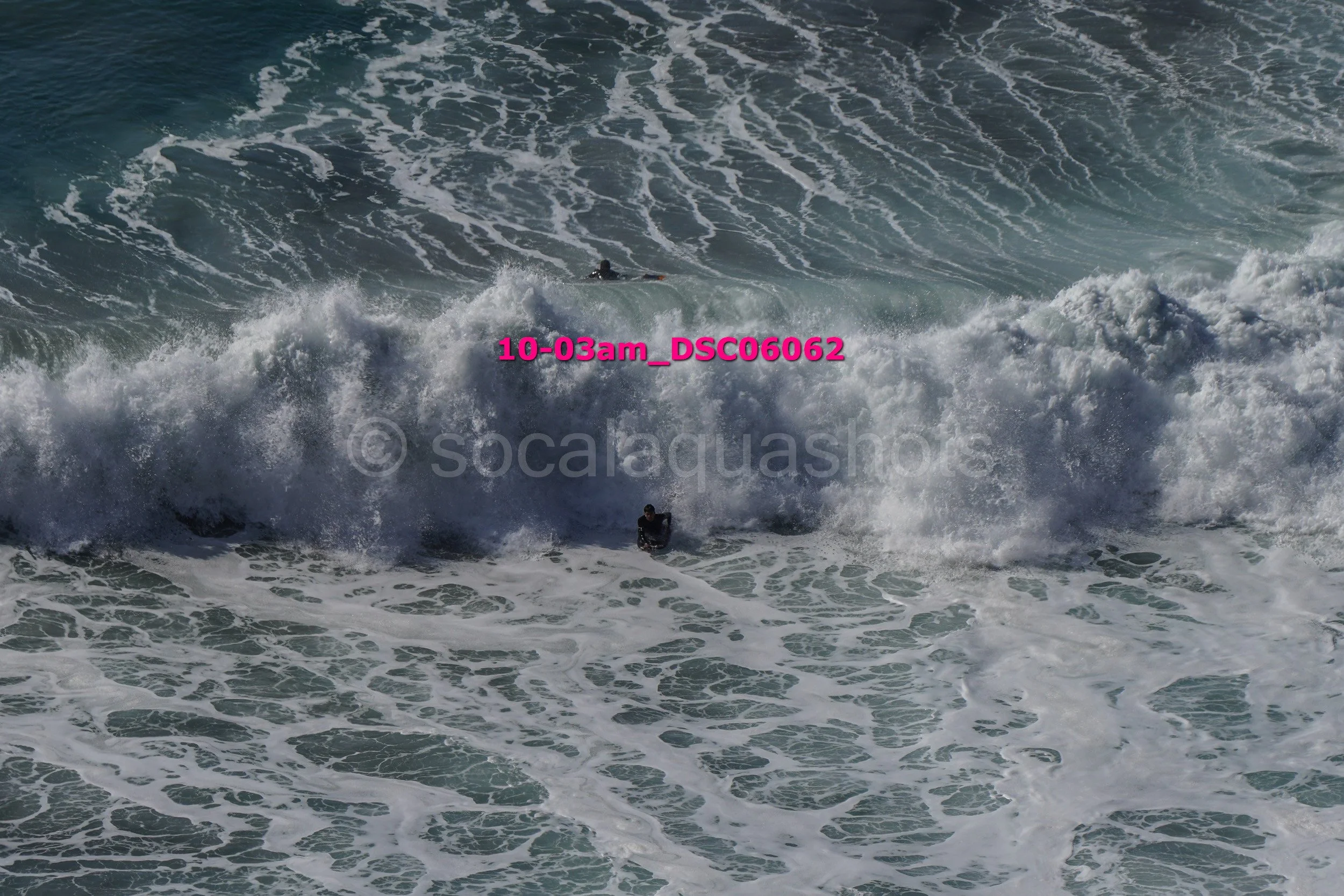 A person surfing in the ocean with large waves and white foam, with another person visible further out in the water.