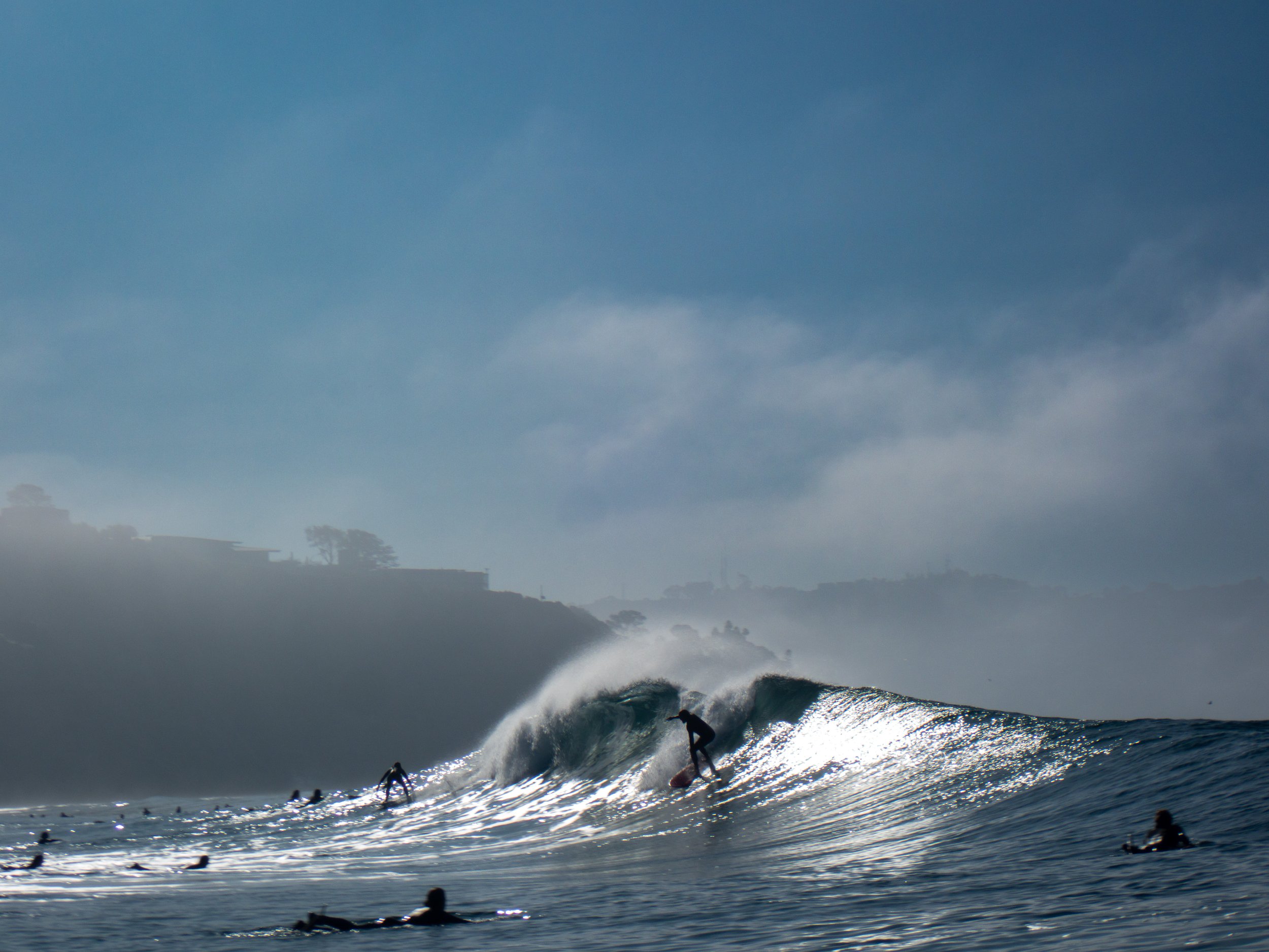Surfers riding a large wave in the ocean with other surfers waiting nearby, against a backdrop of foggy hills and cloudy sky.