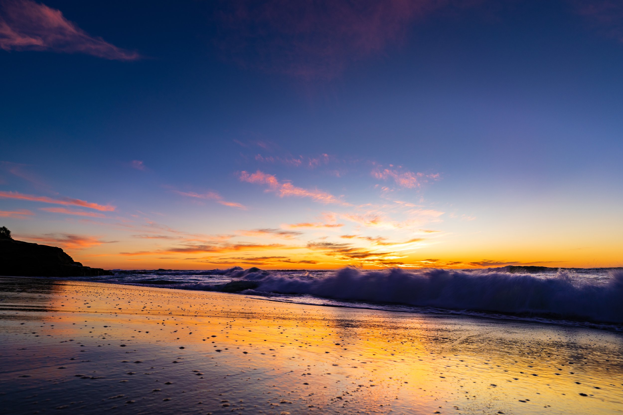 Sunset over the ocean with colorful sky, waves crashing on shore, and wet sand reflecting the sky.