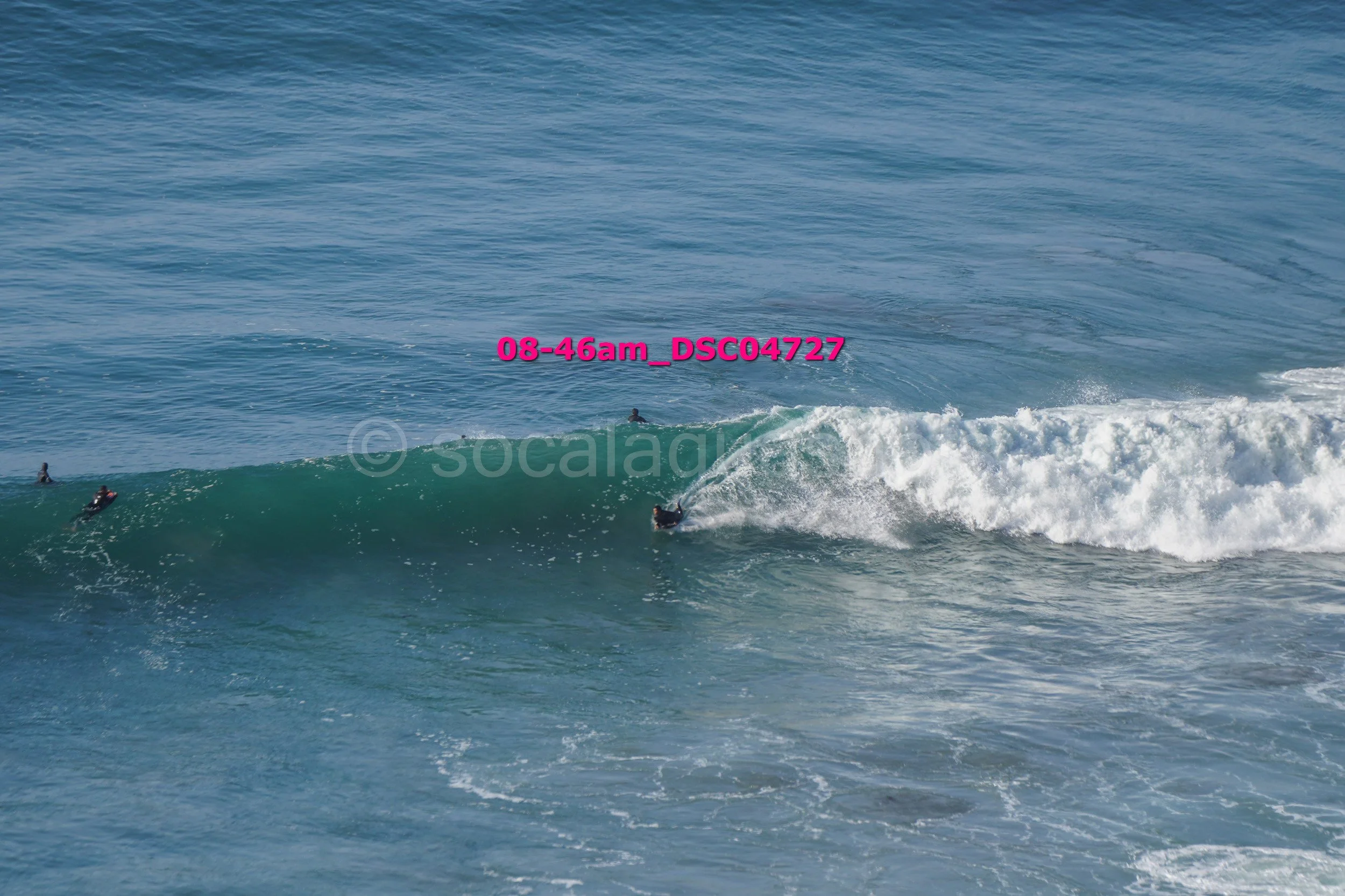 Surfer riding a wave in the ocean with other surfers visible nearby.