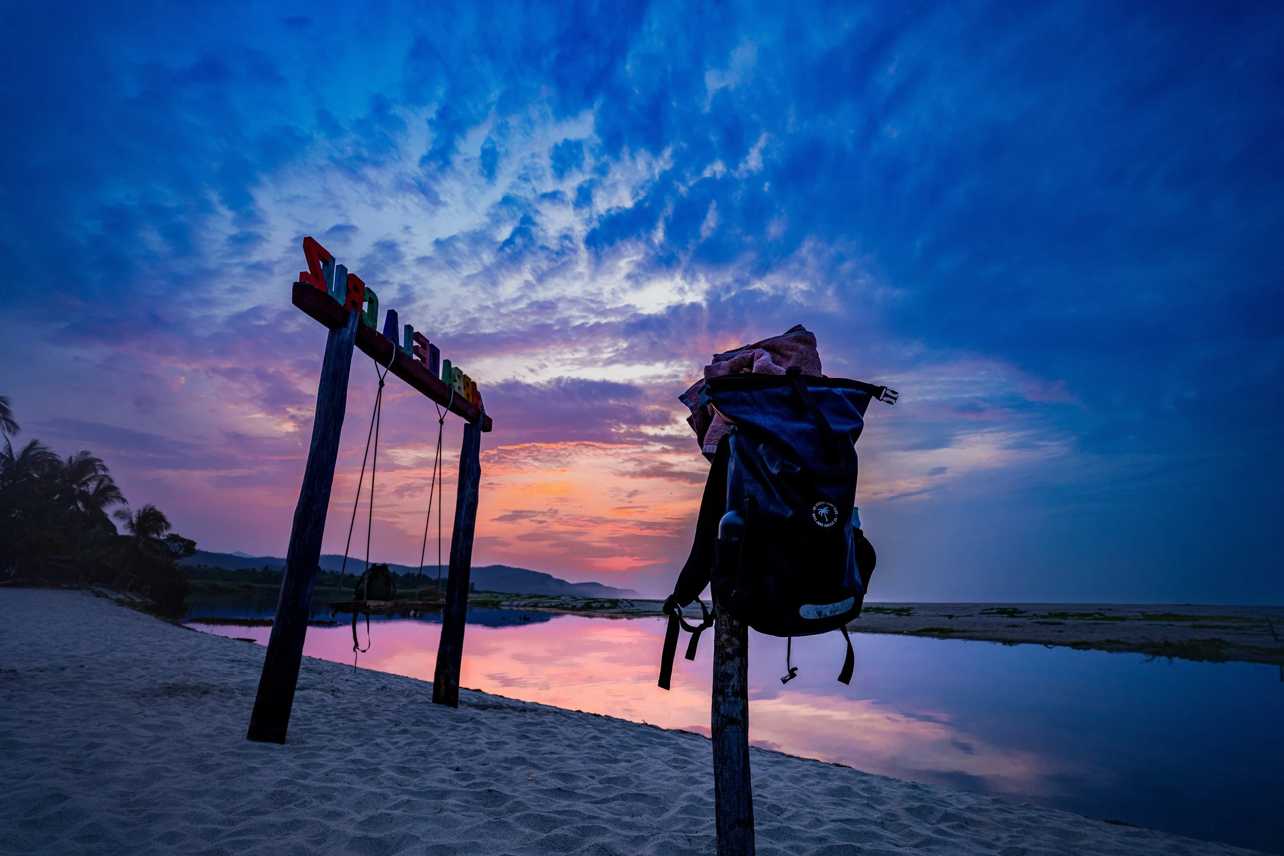 Beach scene at sunset with a sky filled with purple and orange hues, a sign that says 'Welcome,' a basket on a chair, and palm trees in the background.
