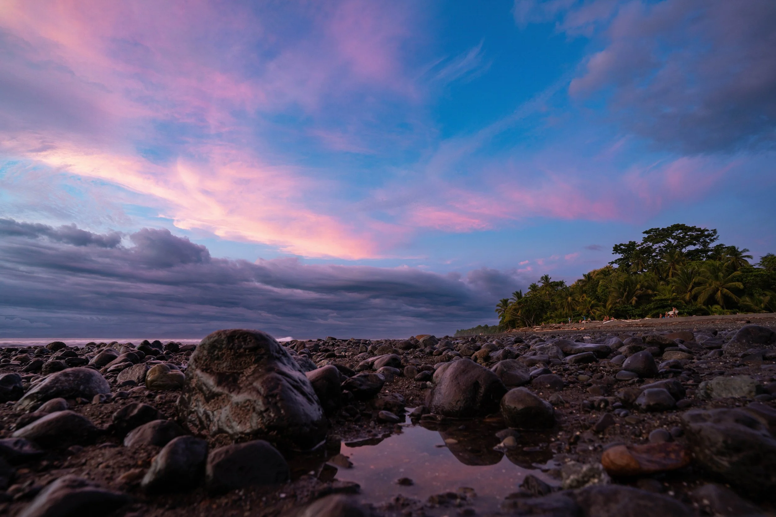 A colorful sunset sky with pink, purple, and blue clouds over a rocky beach with palm trees in the distance.