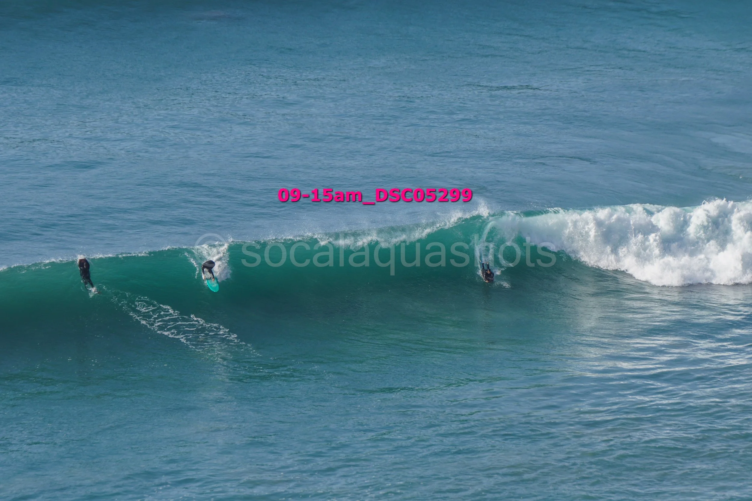 Three surfers riding a large ocean wave with blue water background.