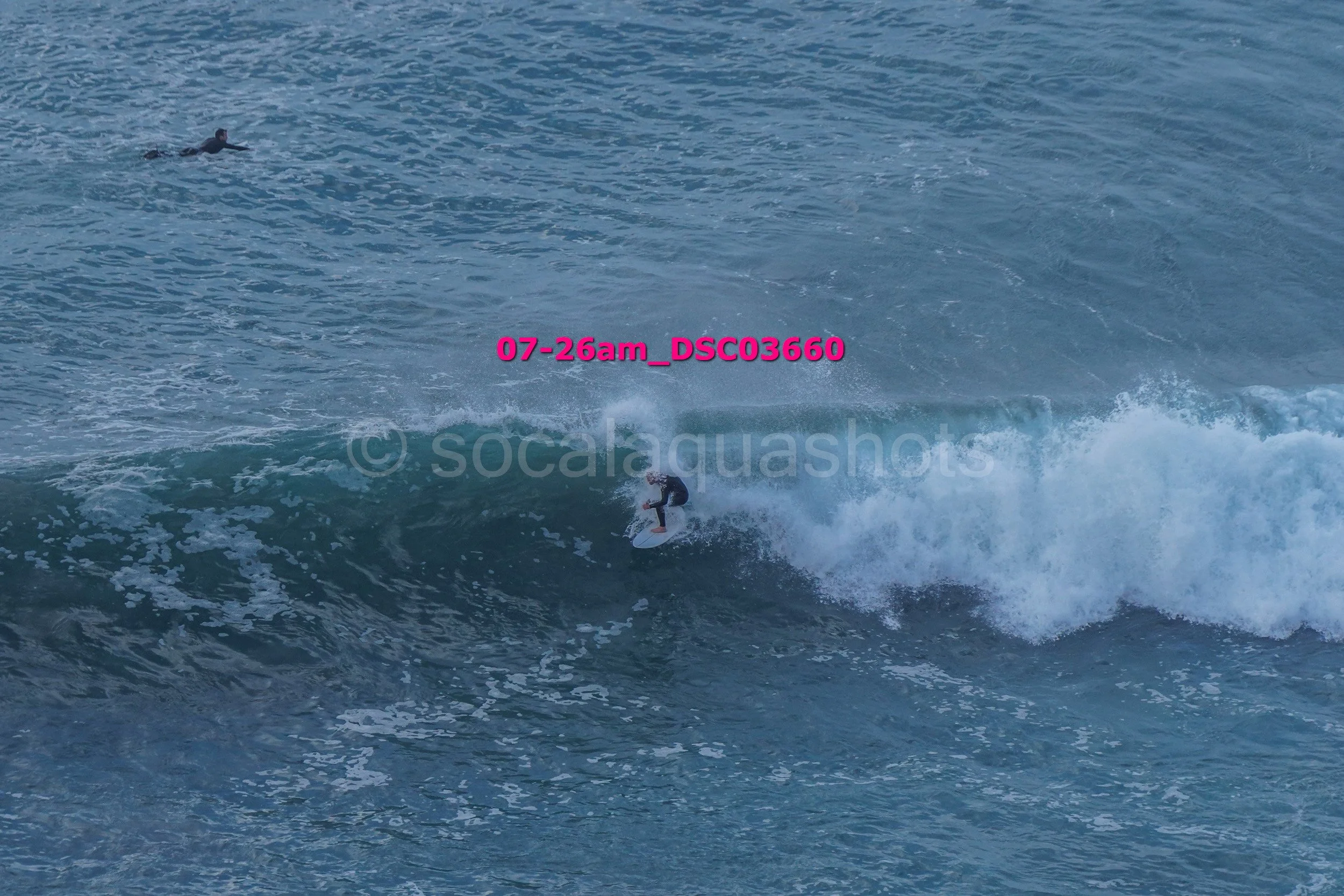 A person surfing on a large wave in the ocean, with another person swimming at a distance.