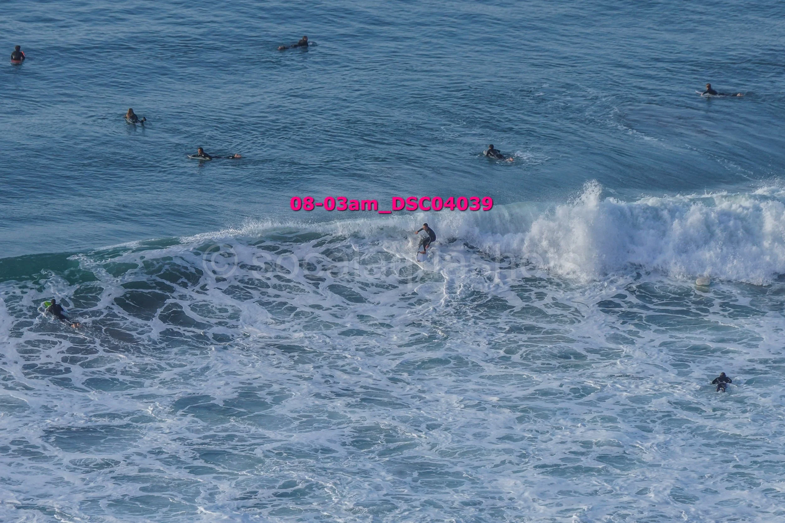 Multiple surfers in the ocean, some riding a wave and others swimming or floating in the water.