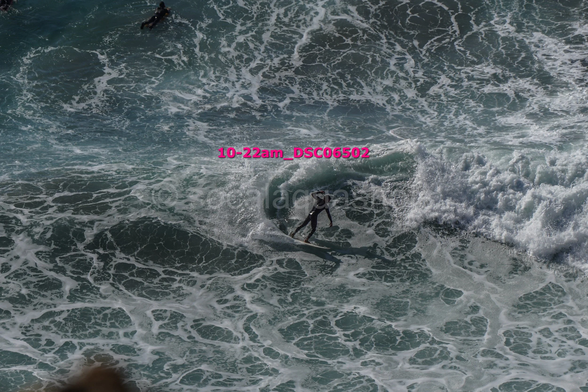 A person surfing on a wave in the ocean during daytime, with another surfer visible further out in the water.