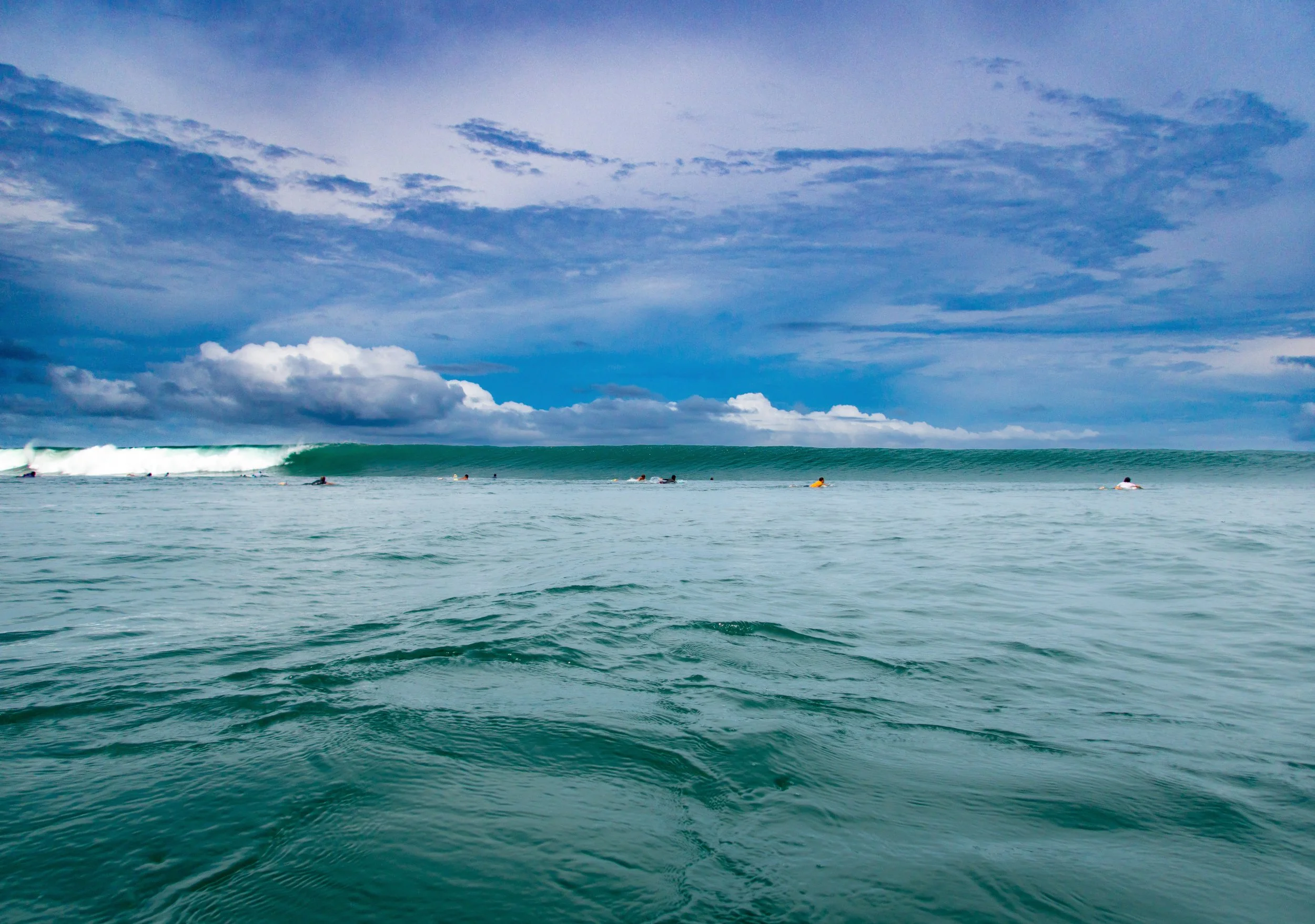 Surfers in the ocean on a cloudy day, sitting on their surfboards near a large wave.