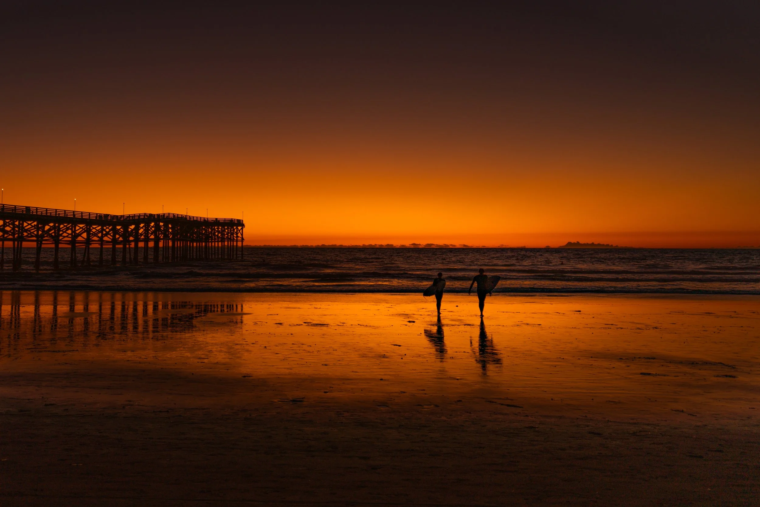 Two surfers walking on a beach at sunset with a pier extending into the ocean on the left side.