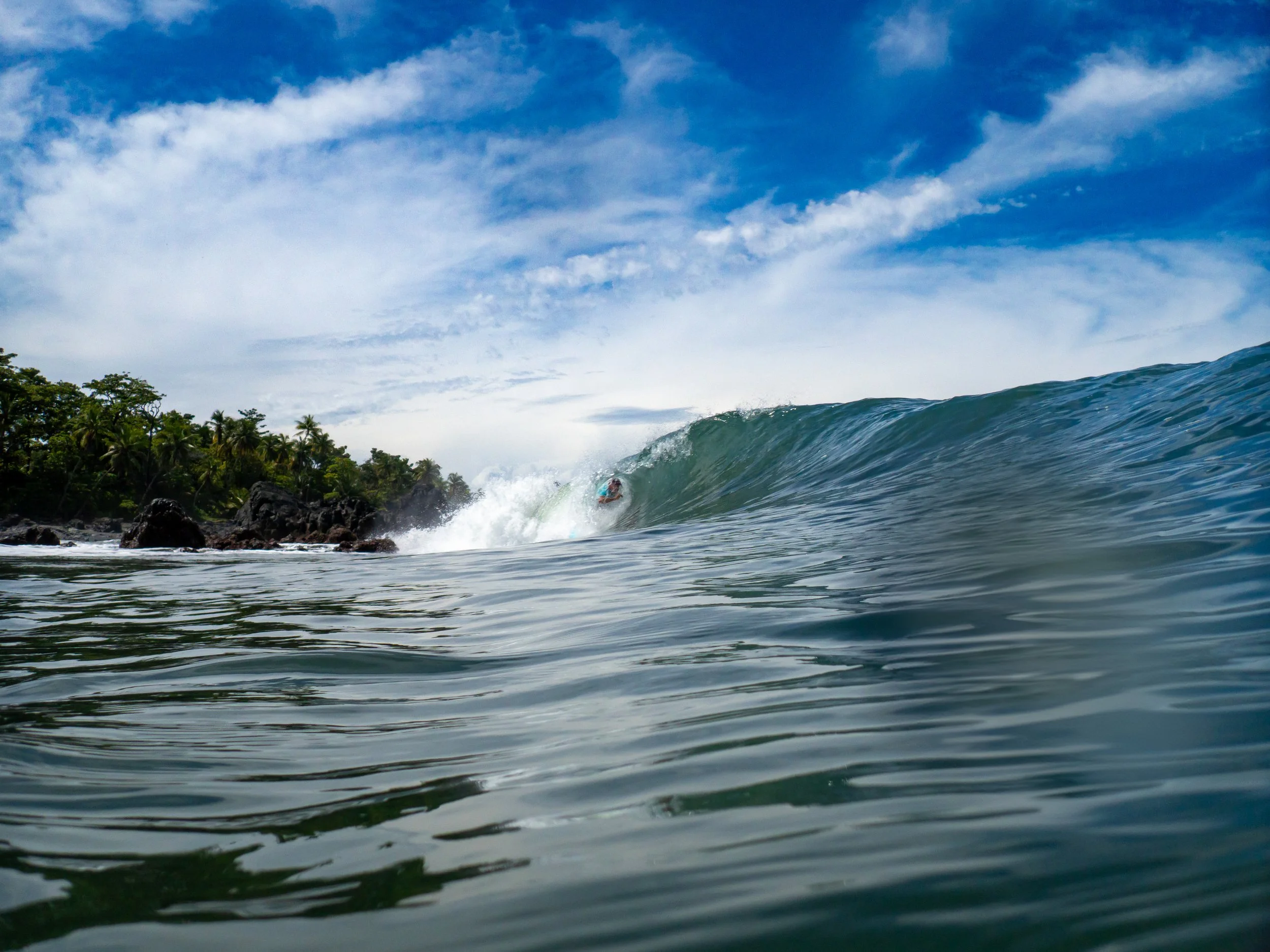 A person surfing on a wave at a tropical beach with lush green trees and rocks, under a partly cloudy blue sky.