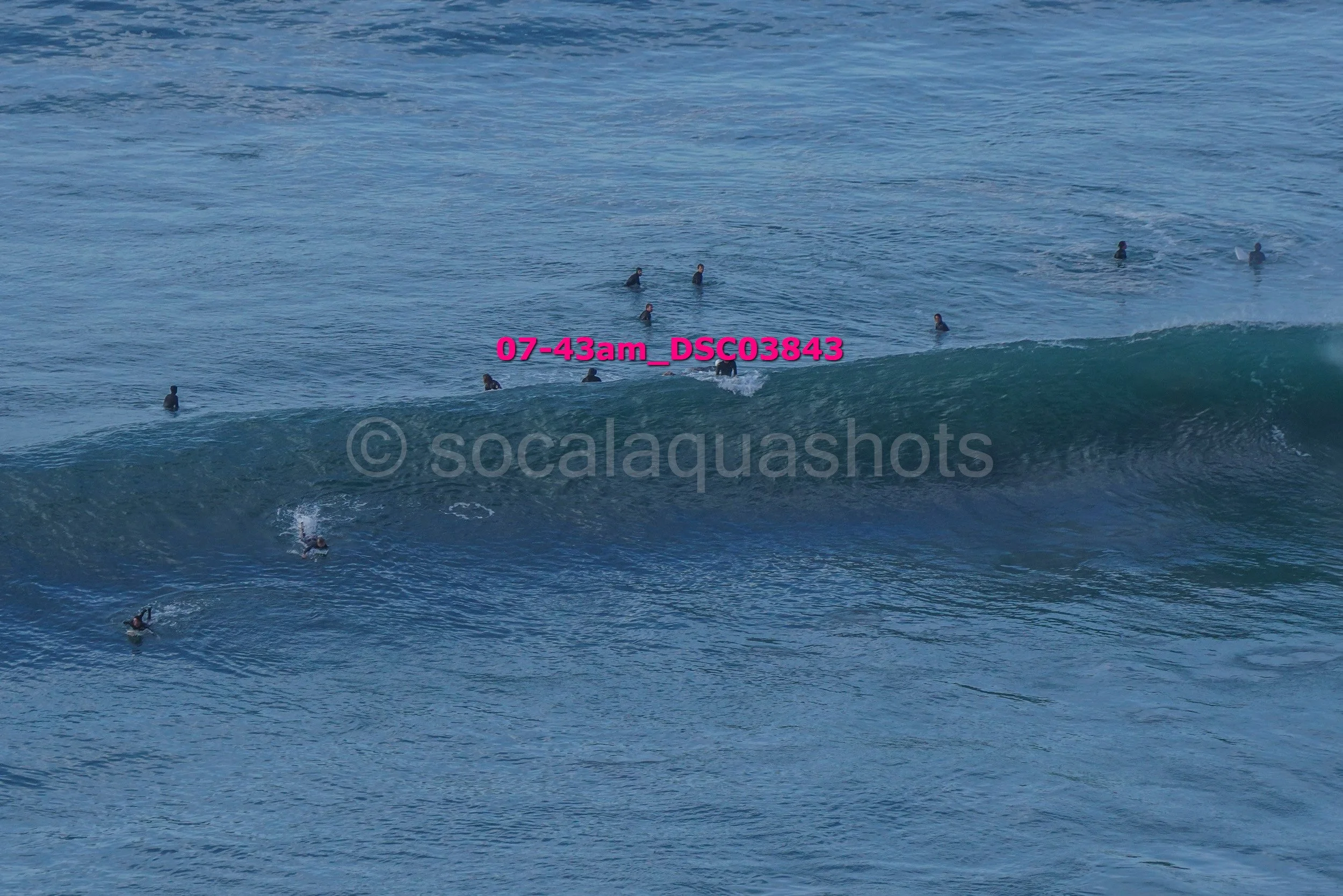 Multiple surfers in the ocean, some riding a wave and others waiting in the water, with a watermark over the image.