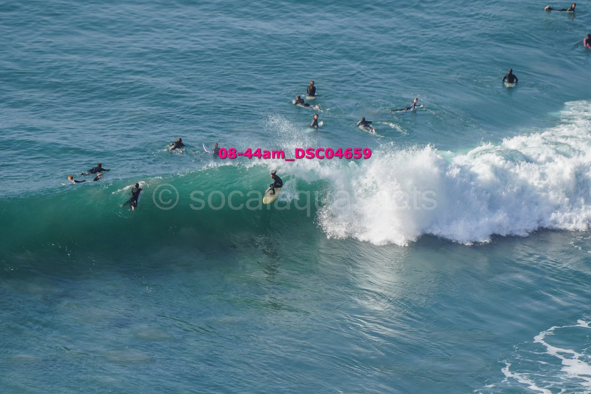 A group of surfers in wetsuits riding and waiting on surfboards in the ocean, with one surfer riding a wave.