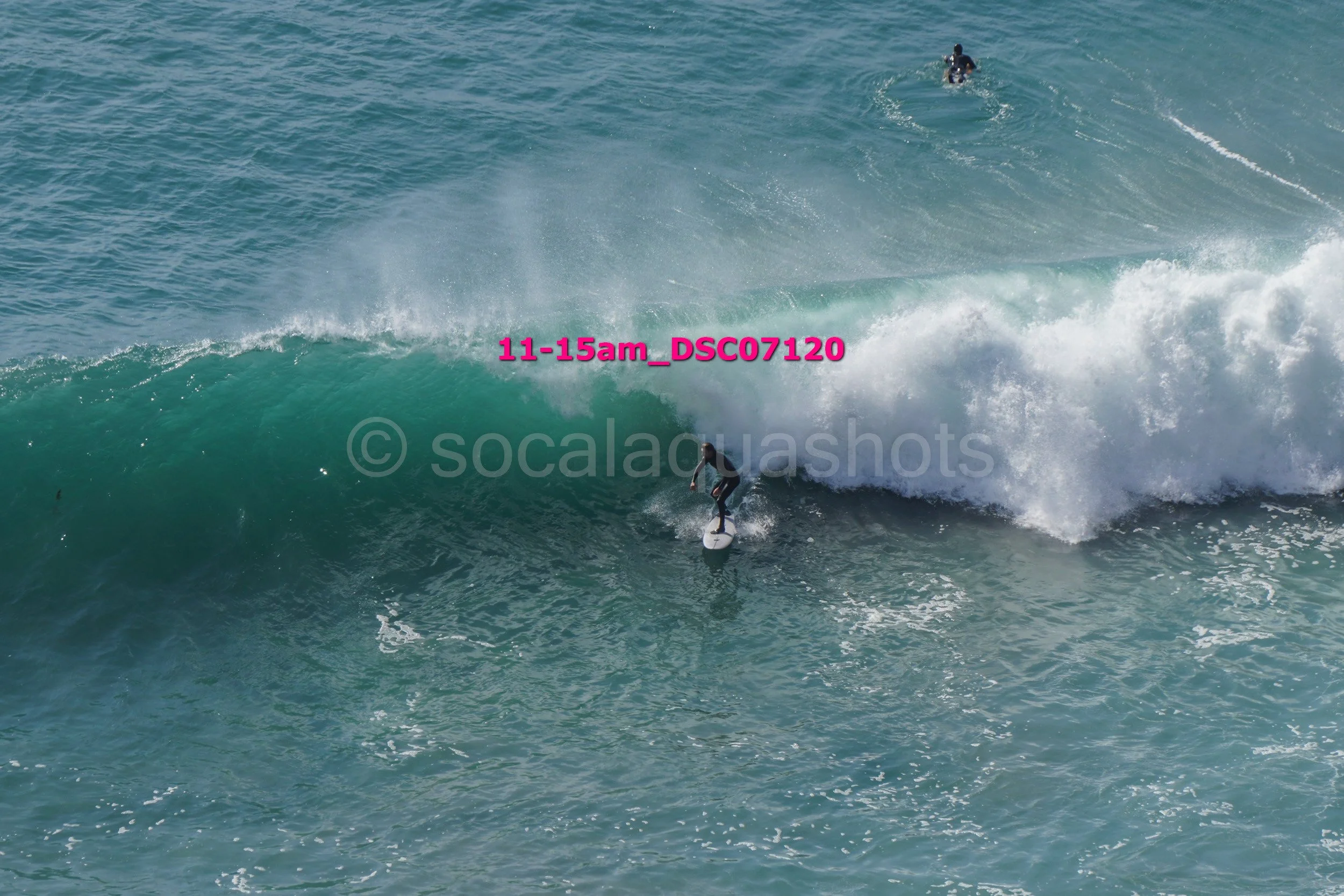 Surfer riding a wave in the ocean with another surfer visible in the background.