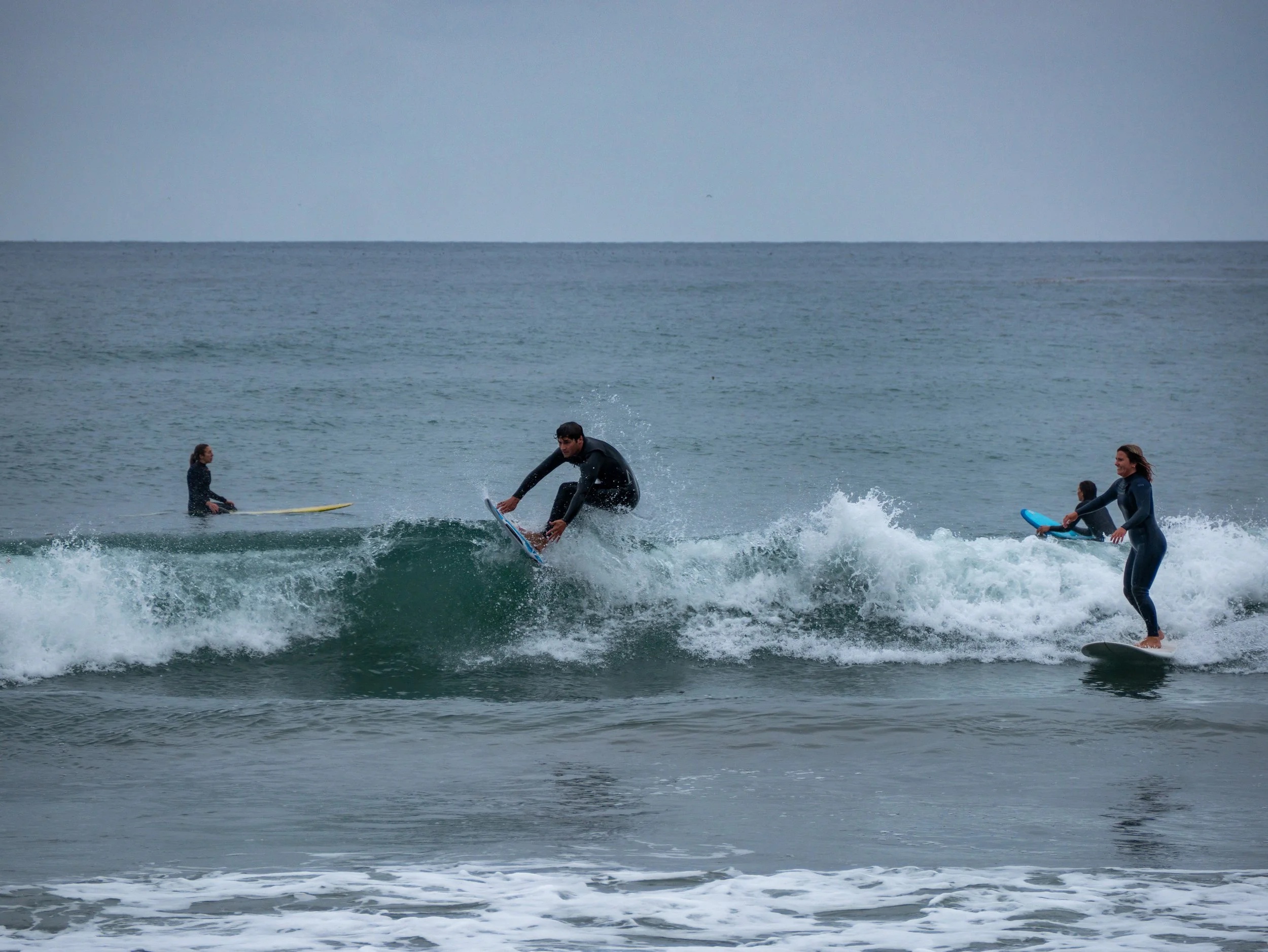 Four people surfing on the ocean, with one person riding a wave and three others waiting on their surfboards.