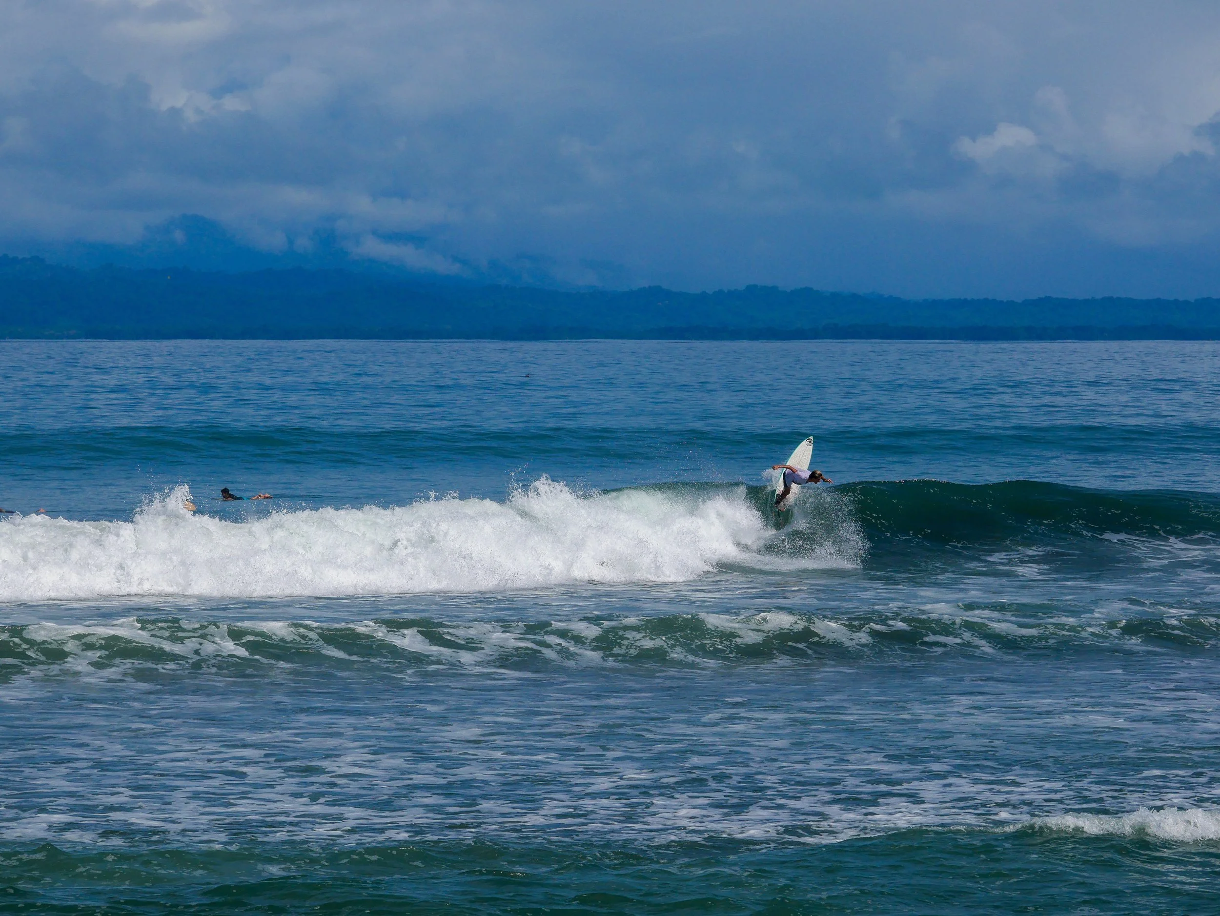 Surfer riding a wave in the ocean under cloudy skies with distant coastline.