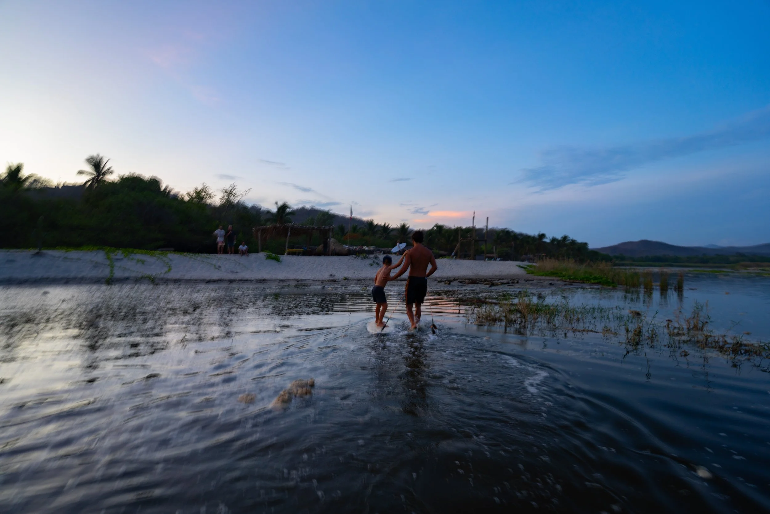 Two children playing in a shallow river at sunset, surrounded by trees and distant hills, with a few people in the background.