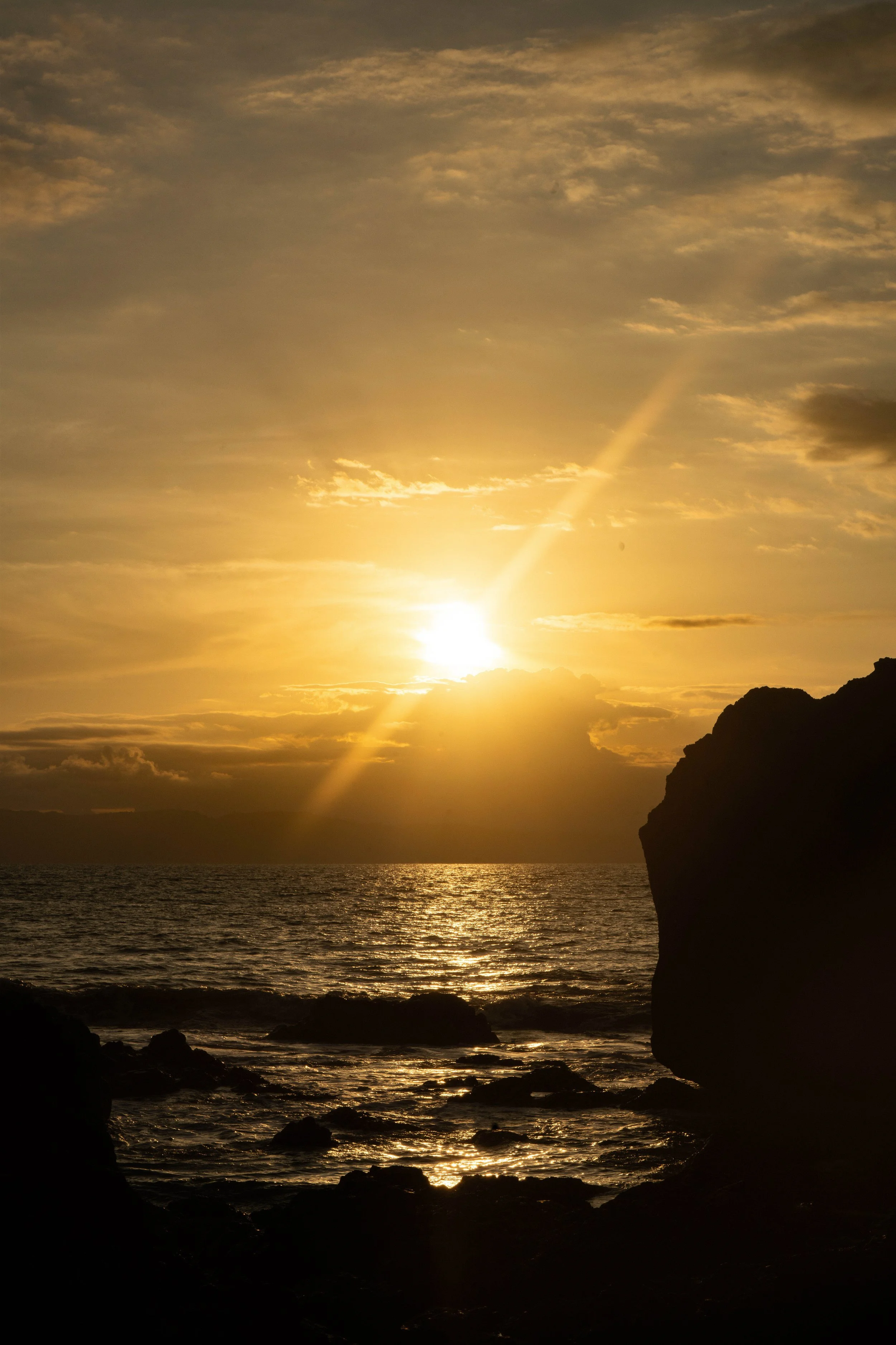 Sunset over the ocean with clouds in the sky, silhouetted rocks along the shoreline, and reflections on the water.