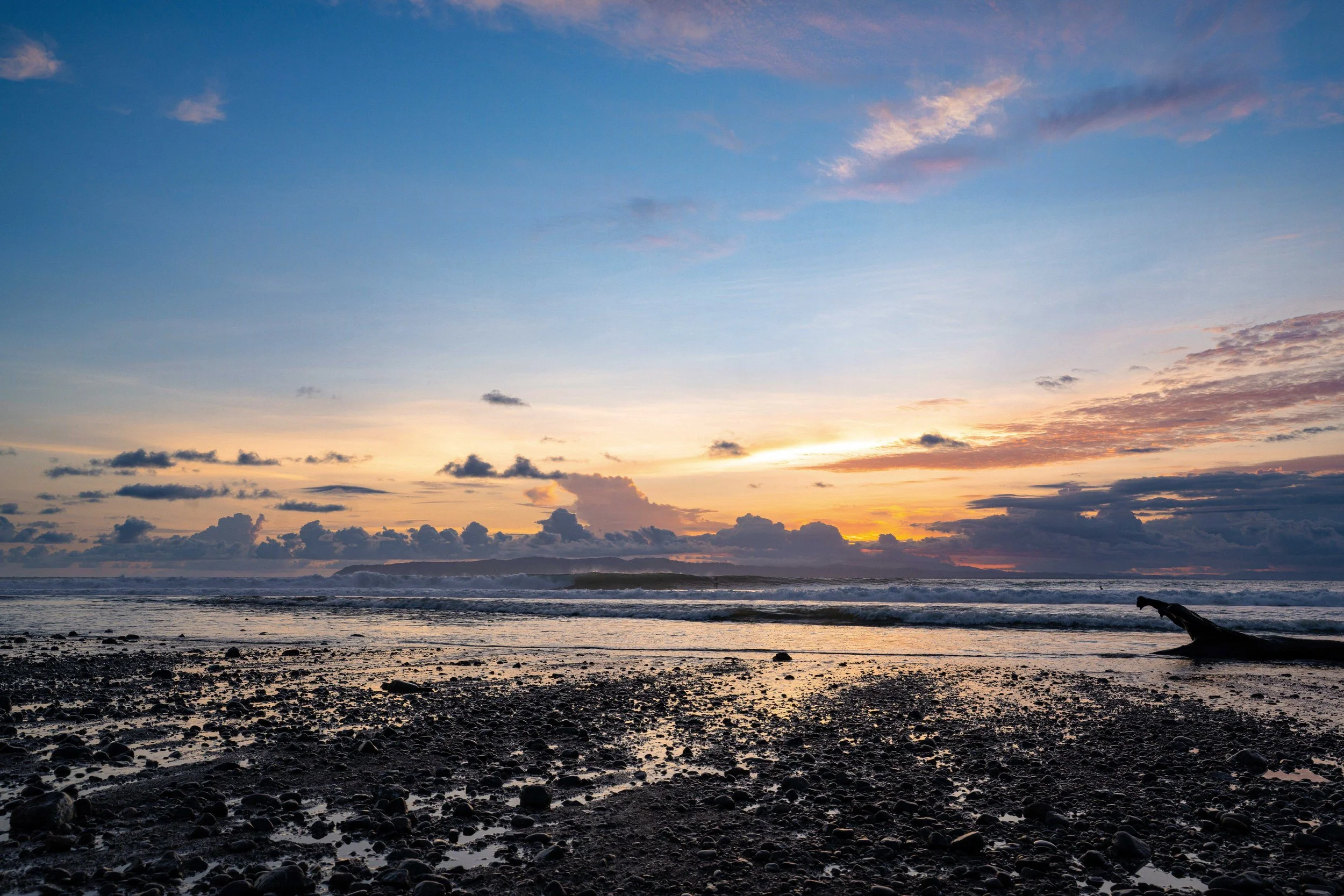 A beach at sunset with a partly cloudy sky, dark rocks and pebbles along the shore, ocean waves, and a distant landform on the horizon.