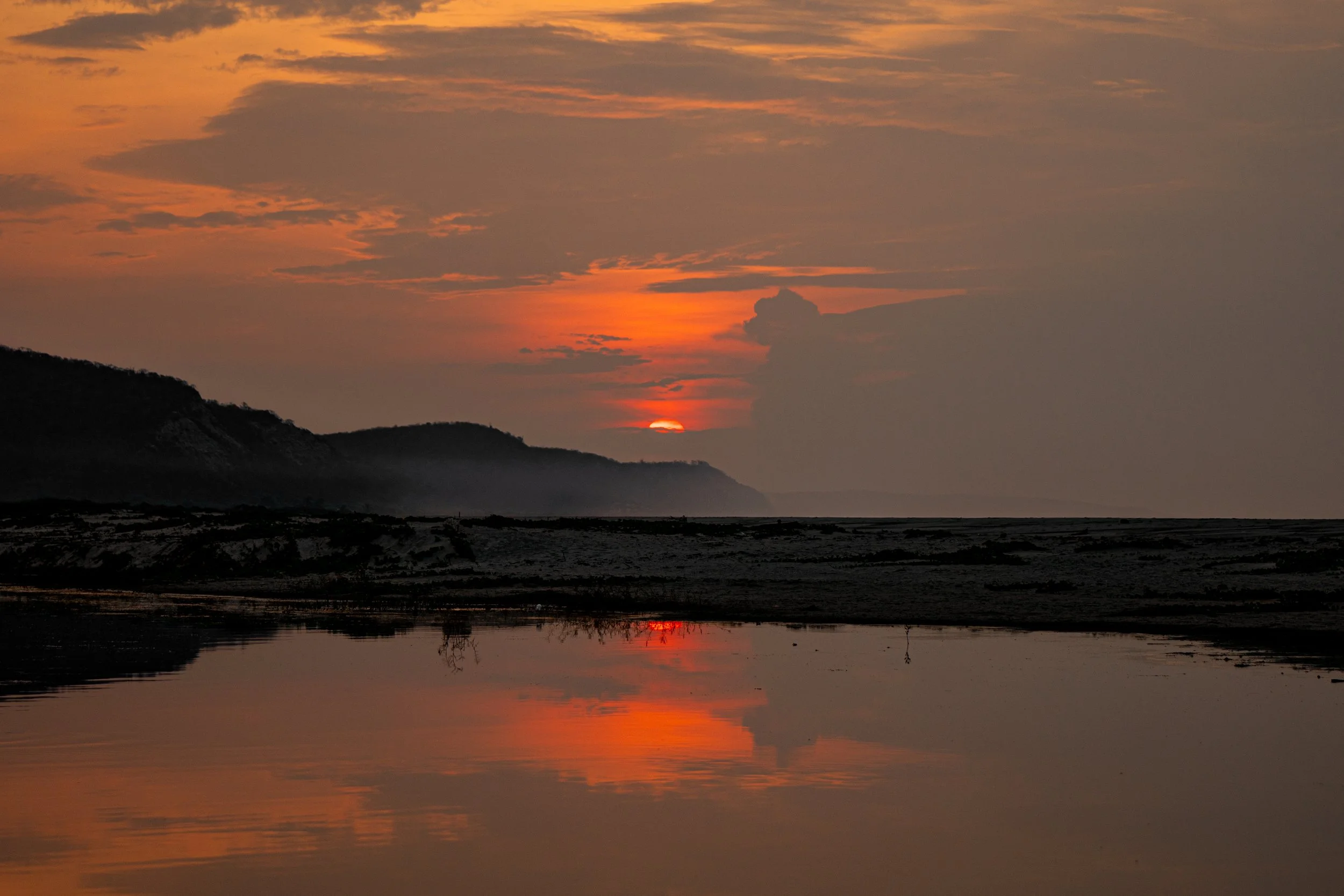 A sunset over mountains with a partly cloudy sky reflected in a body of water.