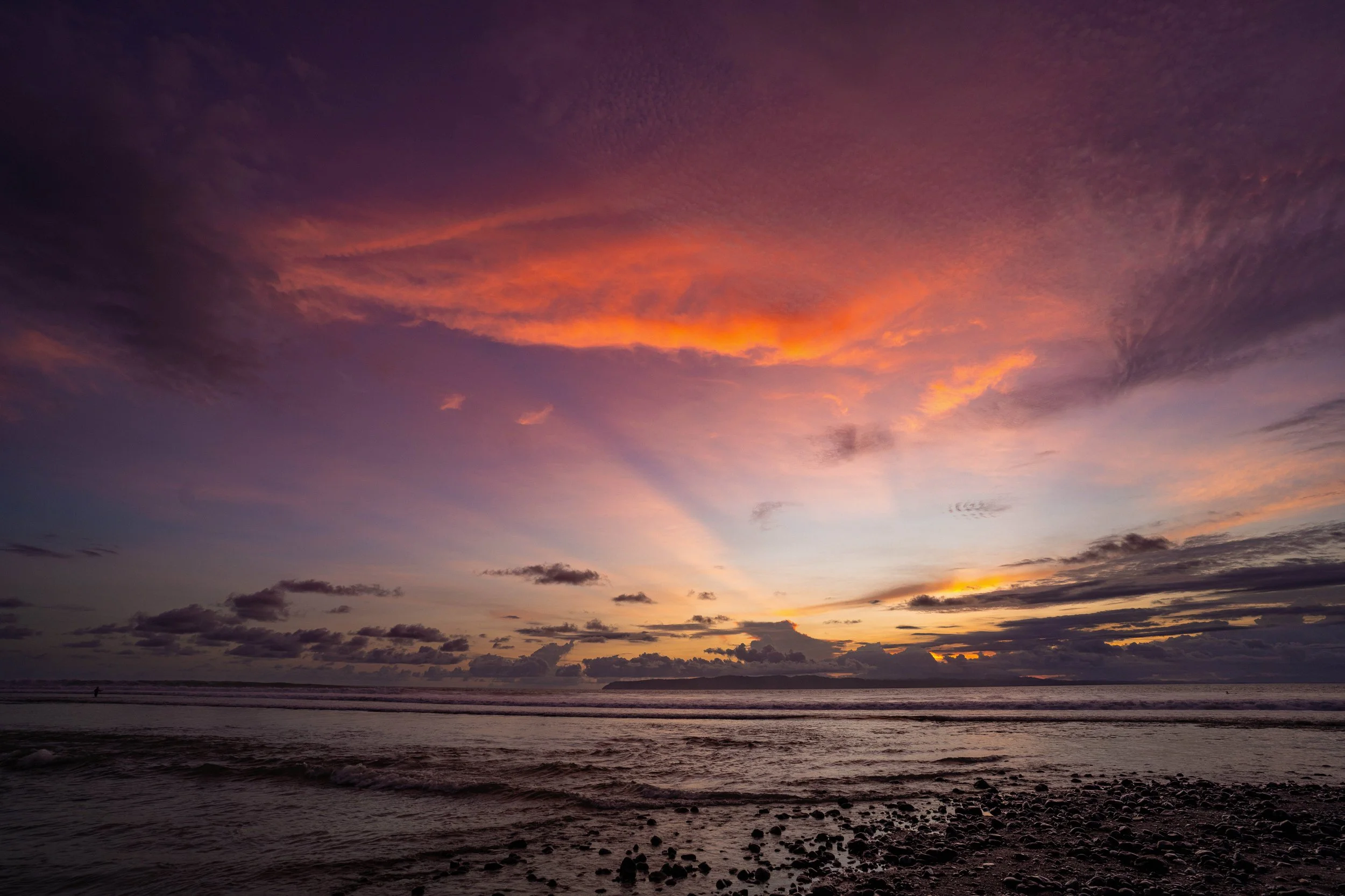 Sunset over the ocean with colorful clouds in the sky, shoreline with rocks and a person walking in the distance.