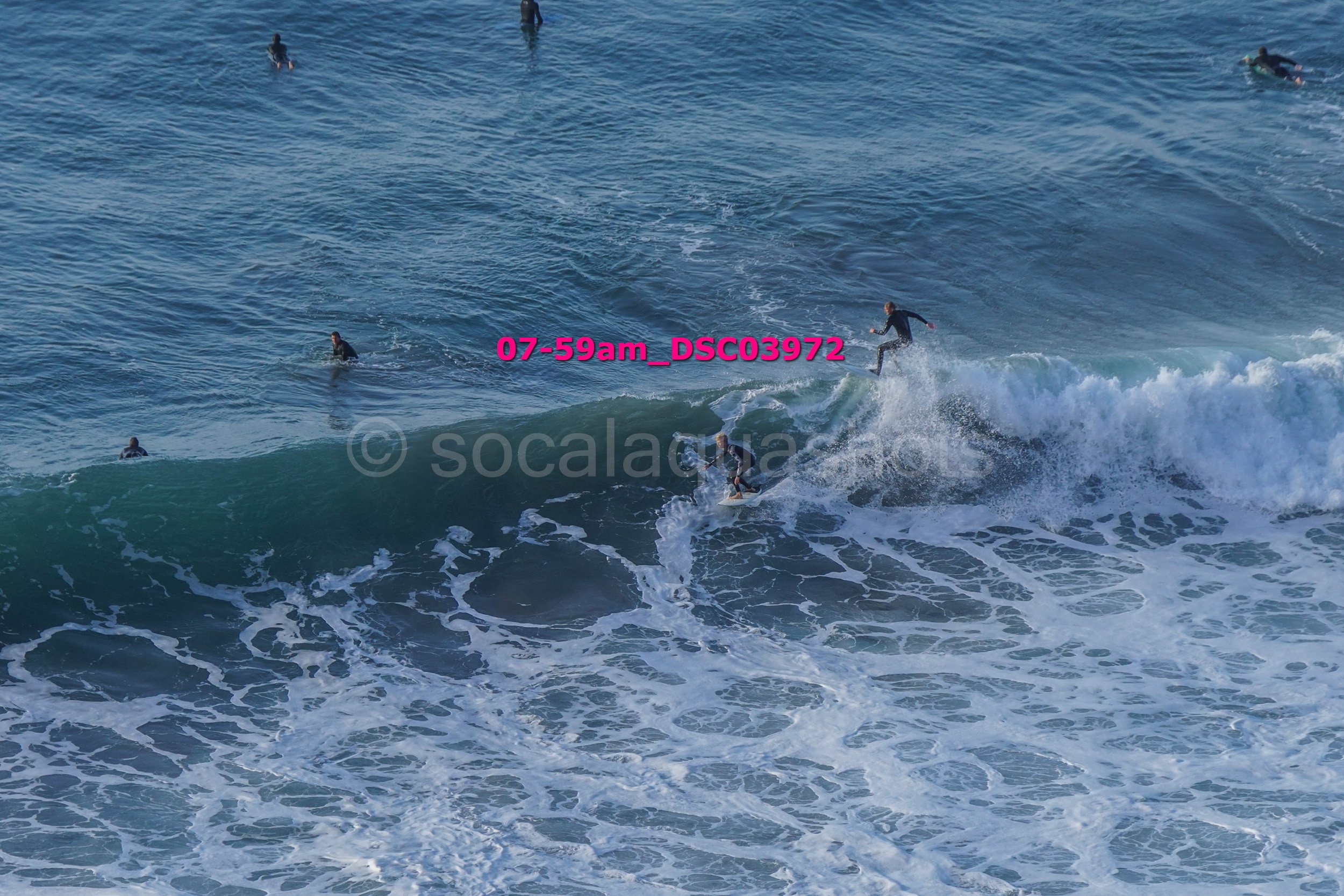 Surfing scene with multiple surfers riding and in the water, large waves crashing, and some surfers waiting in the water.