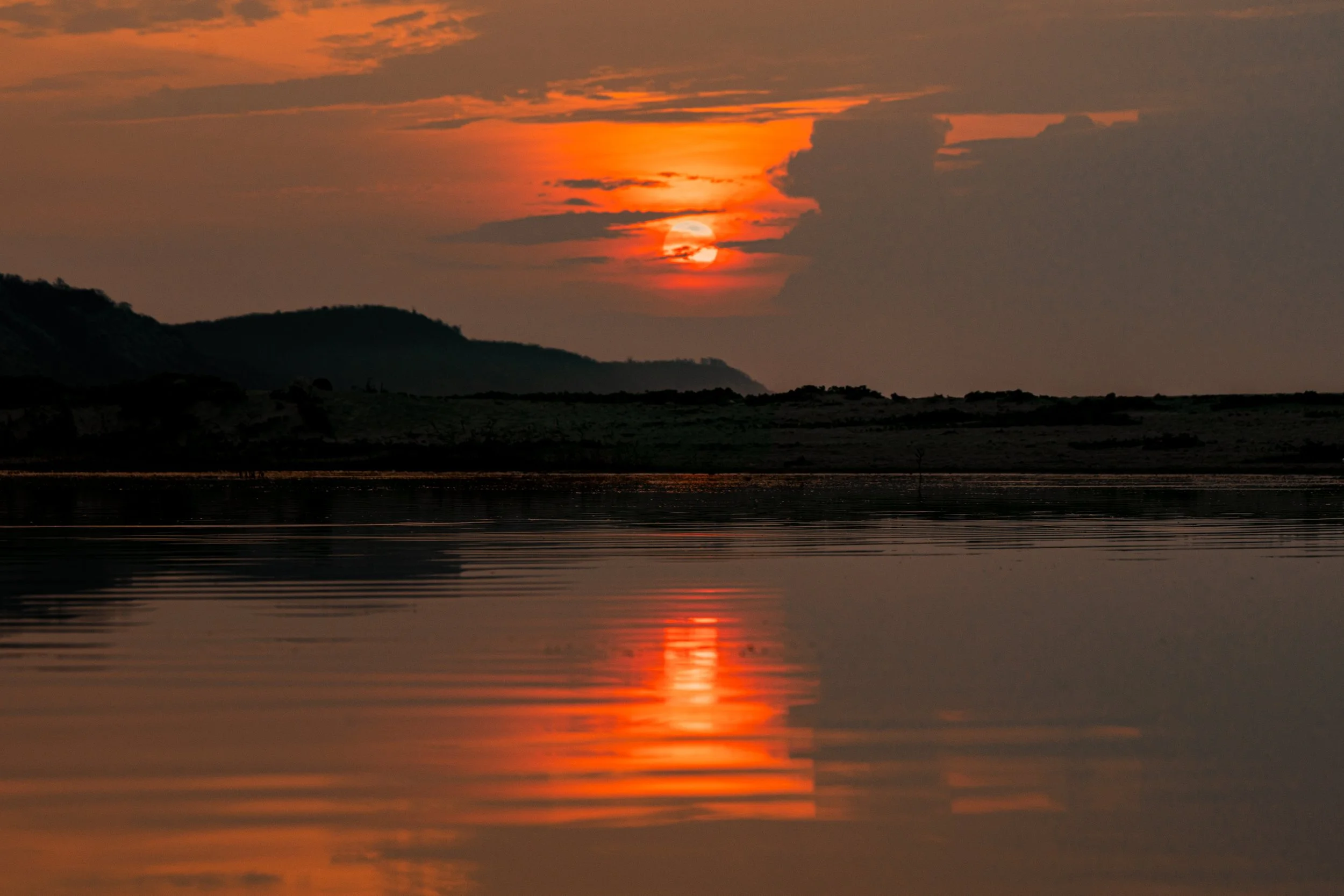 A sunset over a lake, with orange and red hues reflected in the water, silhouettes of distant hills or mountains, and clouds in the sky.