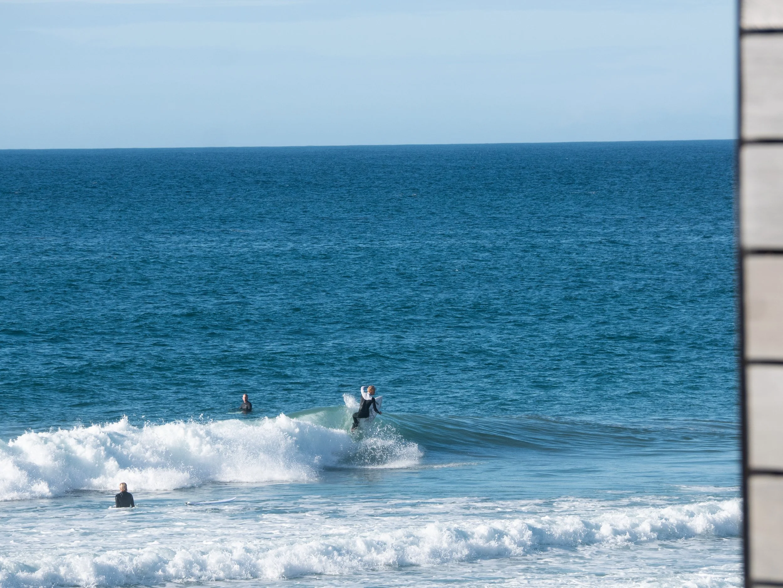 Surfers riding and waiting for waves at the shoreline of the ocean, viewed from beside a wooden structure.