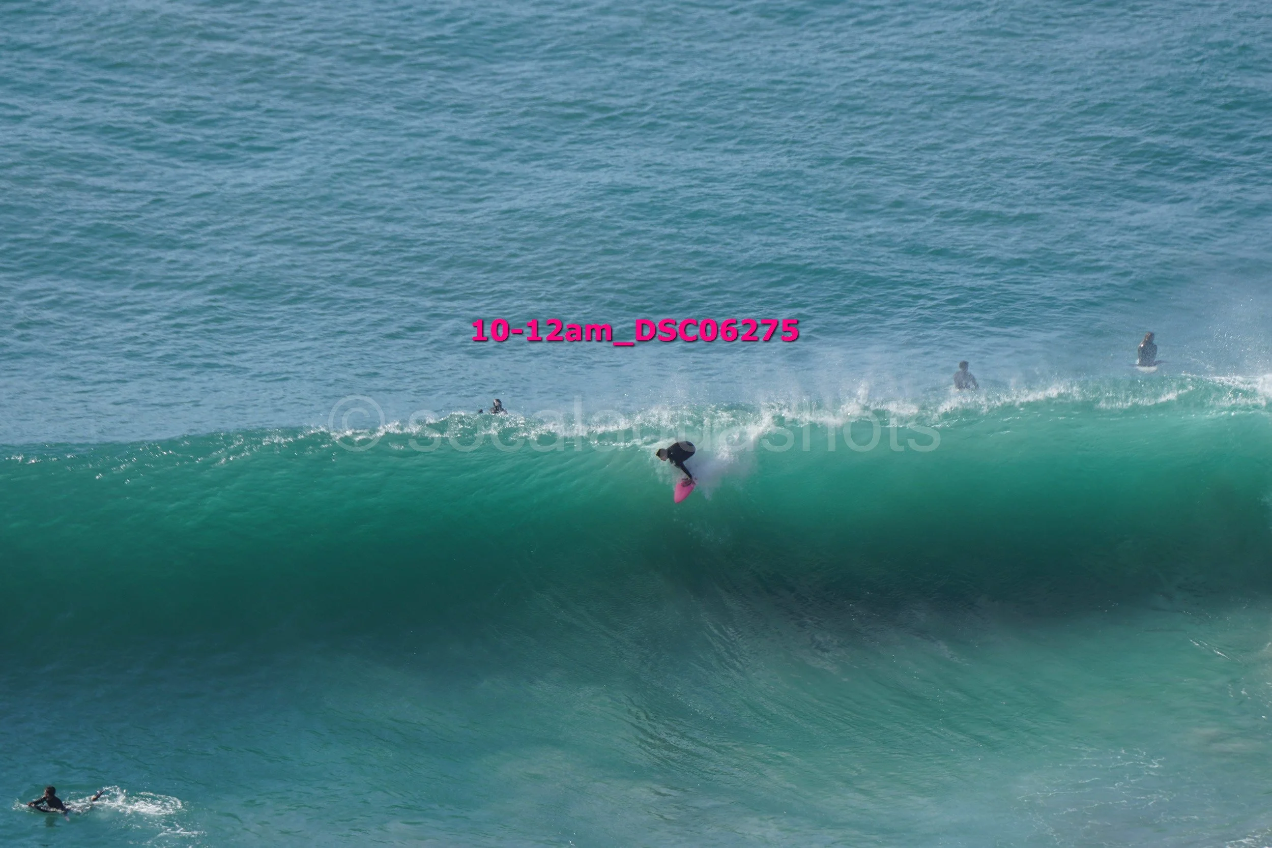 Surfer riding a large wave with other surfers in the background, water splashing, sunny day at the beach.