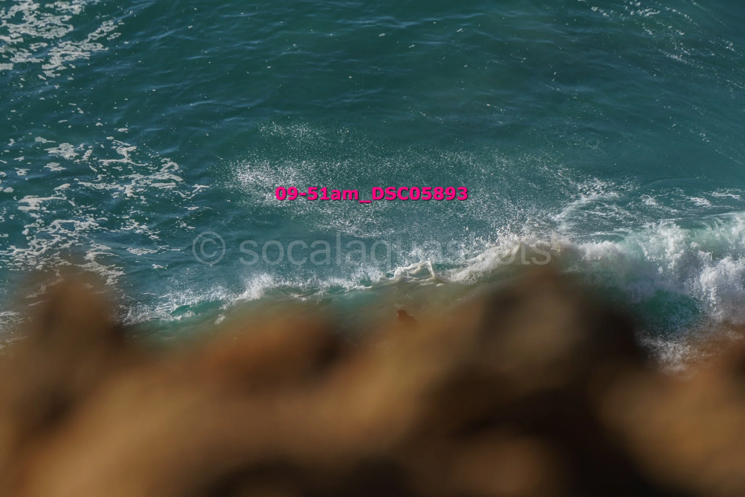 Sea waves crashing against rocks with a person in the water, visible from behind, in the distance.