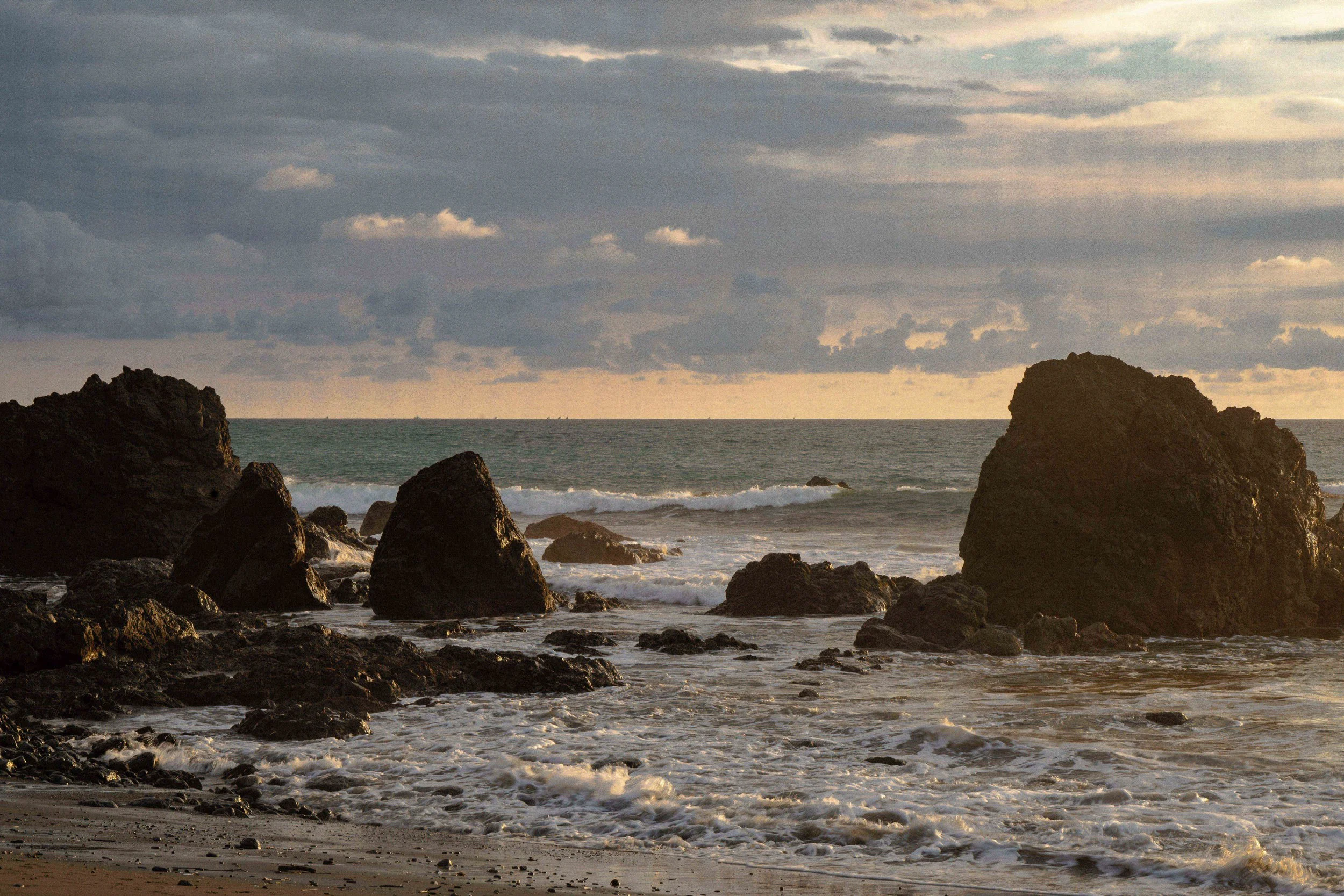 A rocky shoreline at sunset with waves crashing against large rocks and a cloudy sky above.