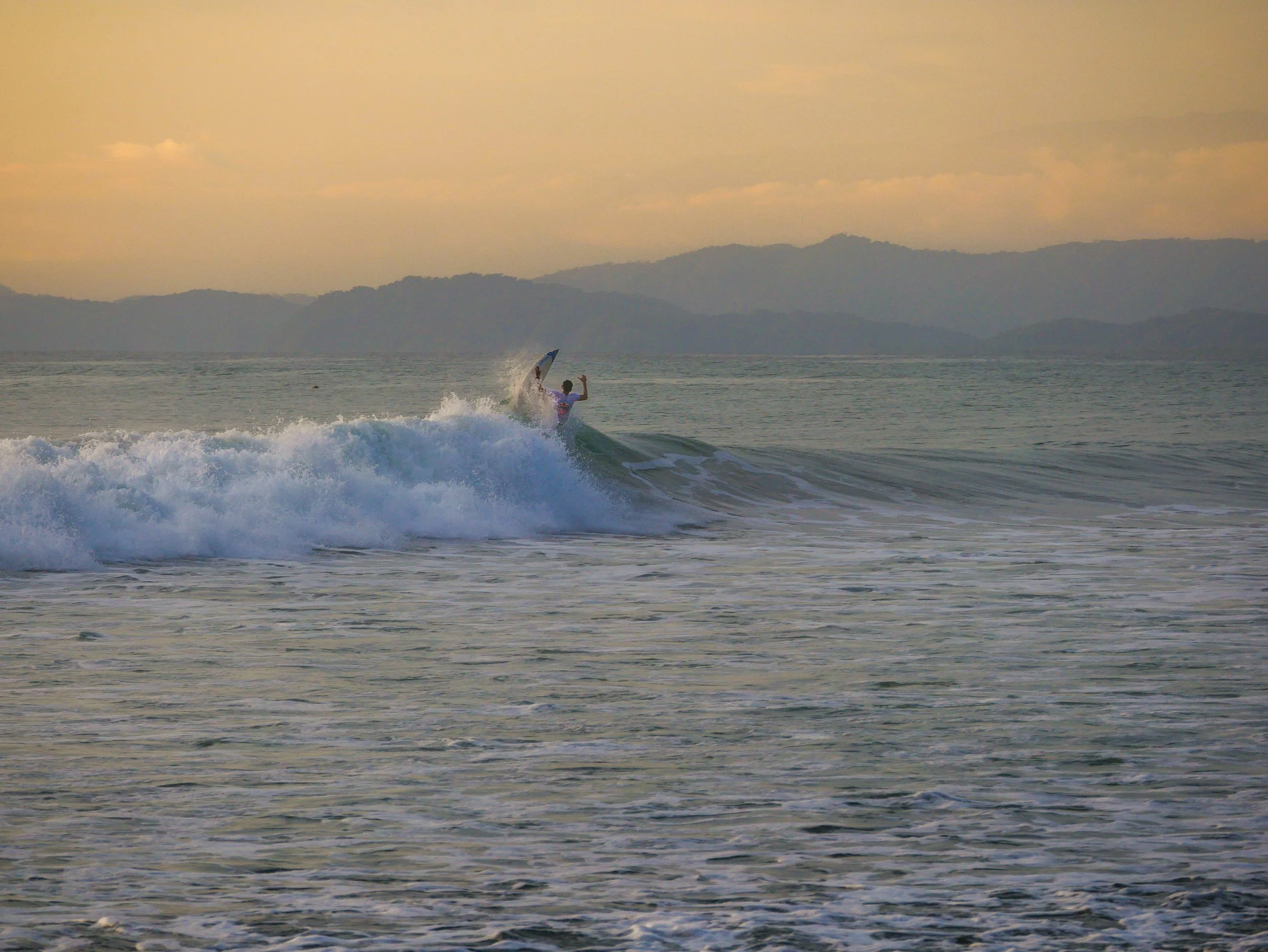 Surfer riding a wave at dawn with mountains in the background.