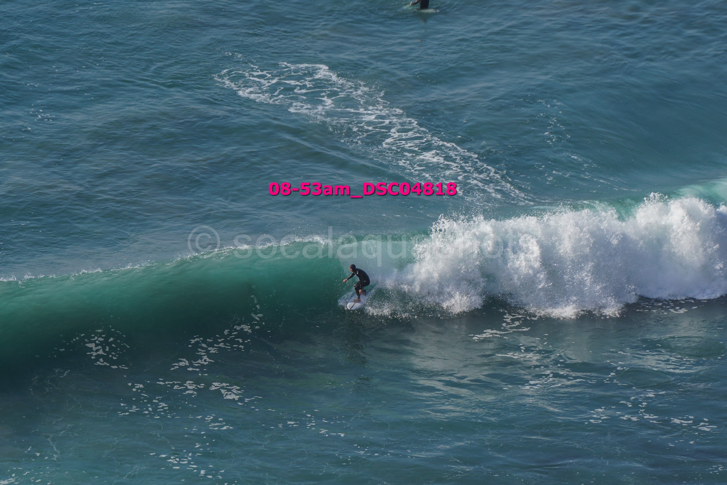 A person surfing on a wave in the ocean, with visible white foam and a deep blue water background.