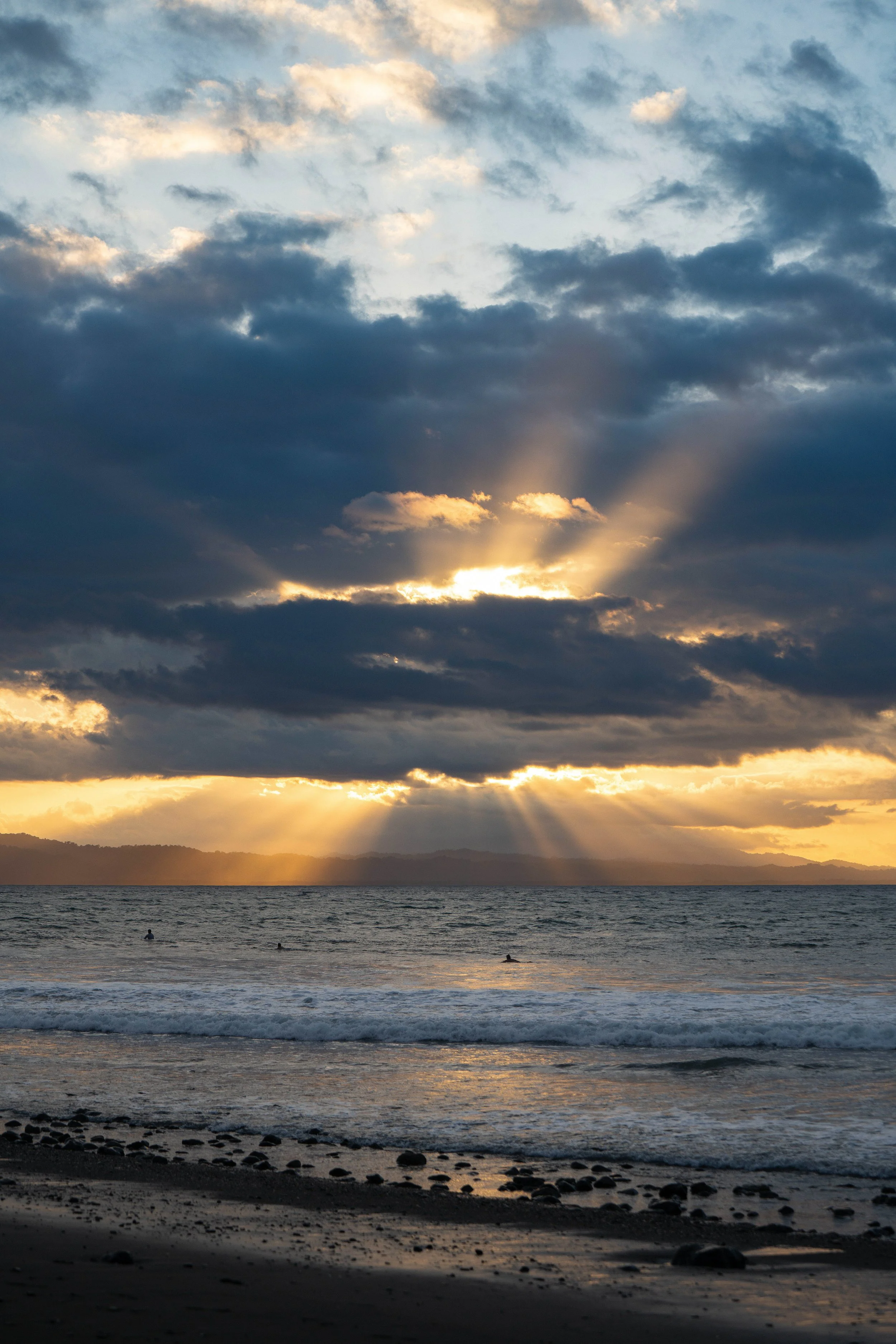Sunset over a beach with dark clouds in the sky, sunlight rays breaking through, calm water, and a rocky shoreline in the foreground.
