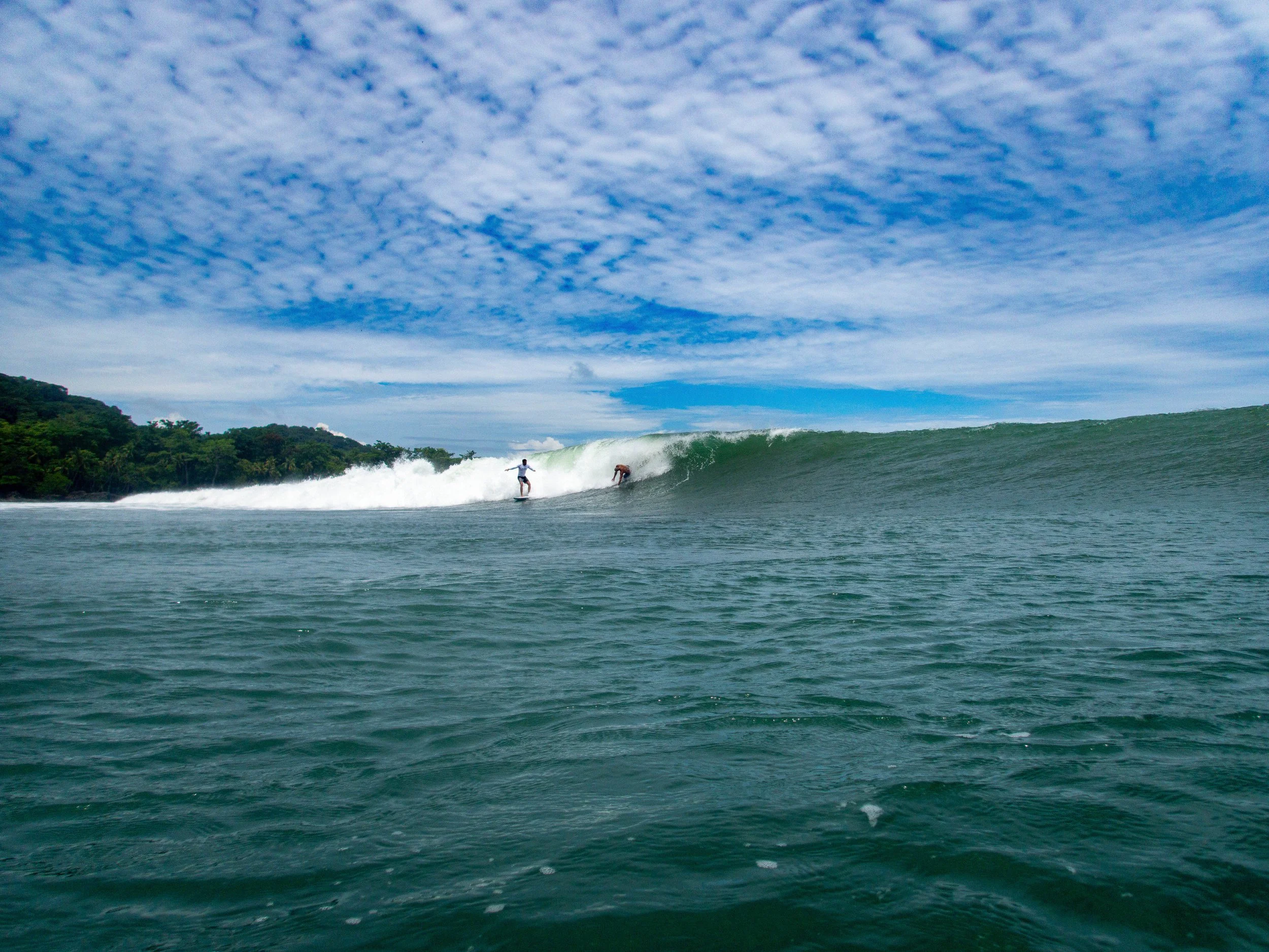 Two surfers riding a large ocean wave under a cloudy blue sky near lush green coastline.