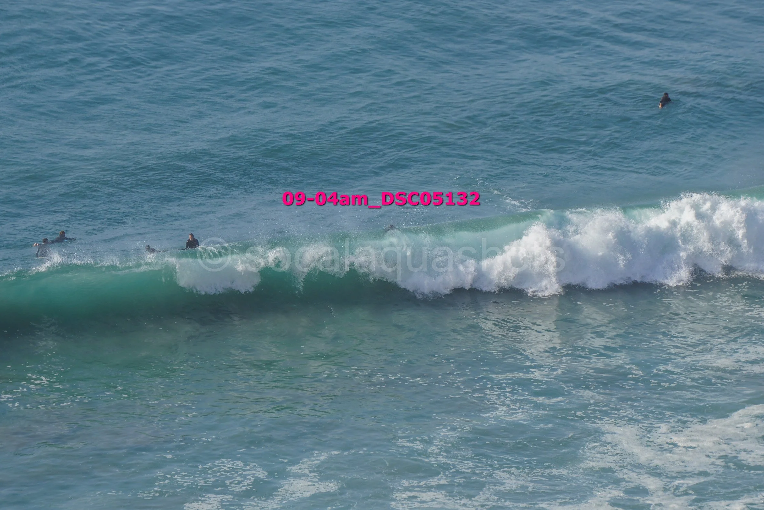 Several people are surfing on the ocean waves, with some lying on surfboards and others standing or paddling in the water.