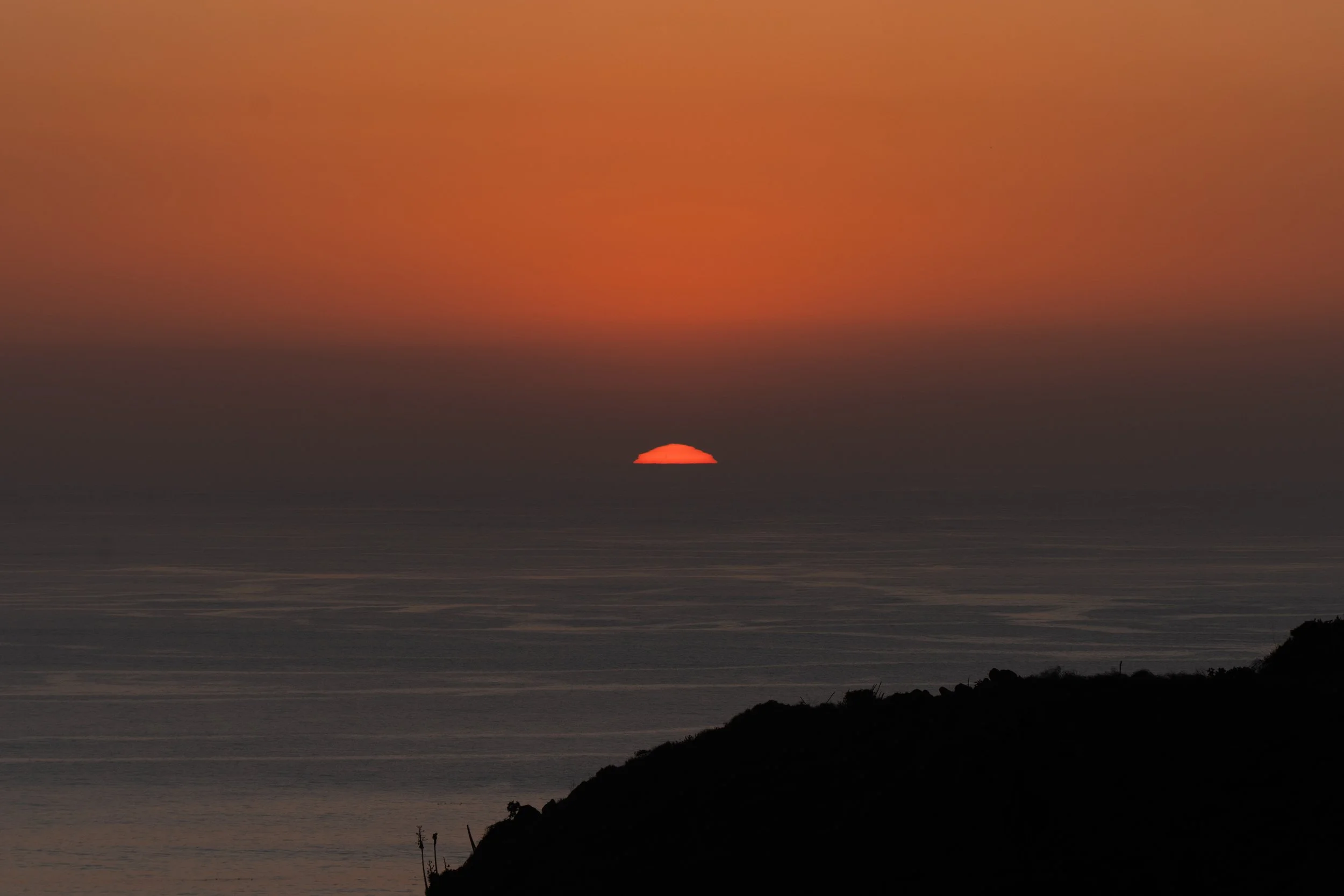 A sunset over the ocean with the sun partially below the horizon and a silhouetted landmass in the foreground.