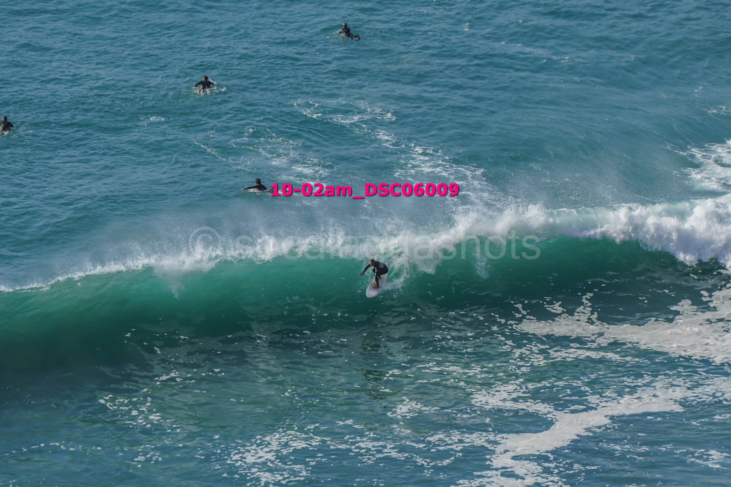 Surfer riding a wave in the ocean with several people swimming and surfing in the background.
